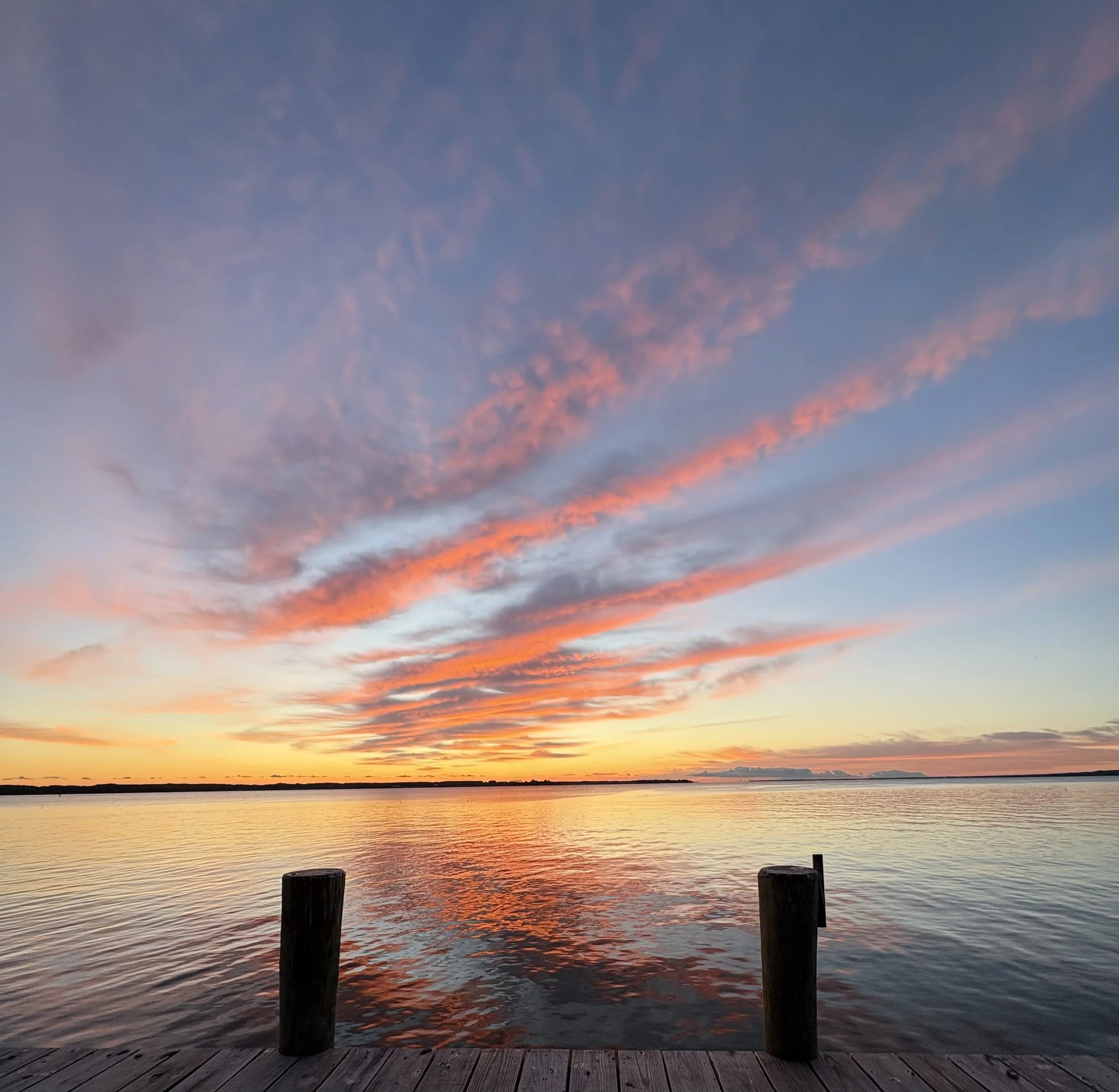 Sunset over a calm body of water, viewed from a wooden pier with fence posts in the foreground.