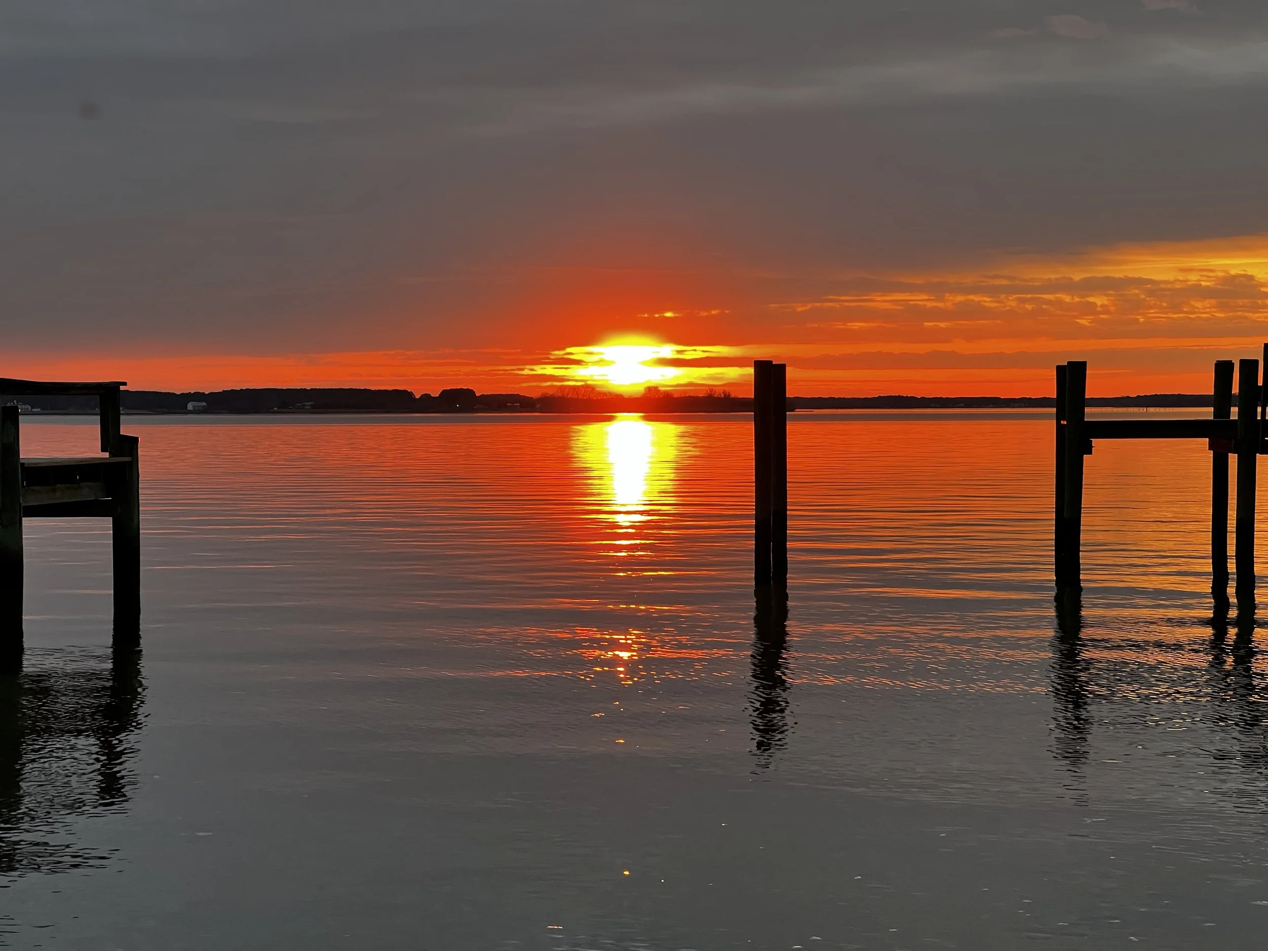 Sunset over a calm body of water with silhouettes of a pier and distant shoreline, reflecting vibrant orange and yellow hues in the sky.