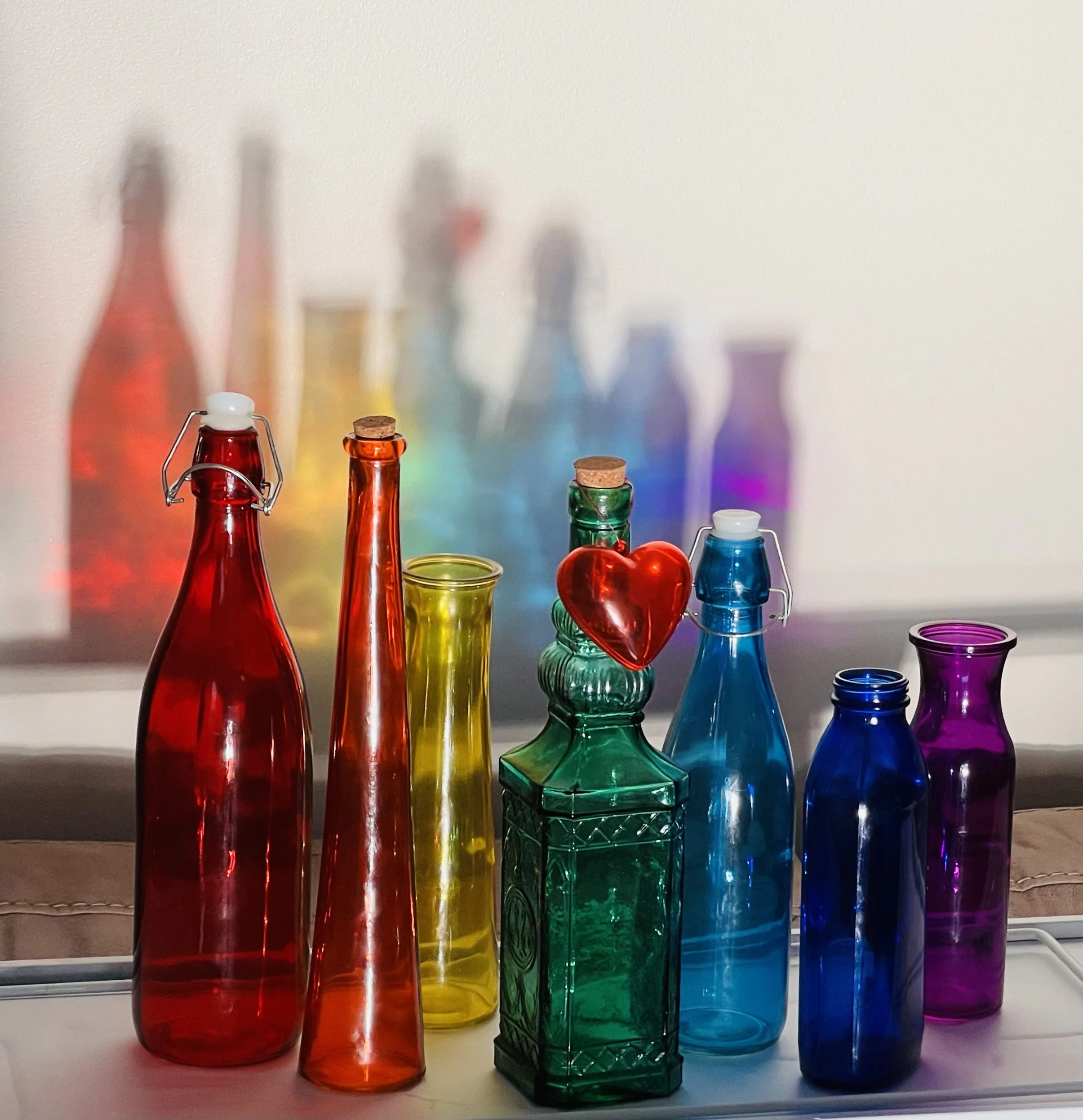 Colorful glass bottles of various shapes and sizes arranged on a shelf with a white wall in the background.
