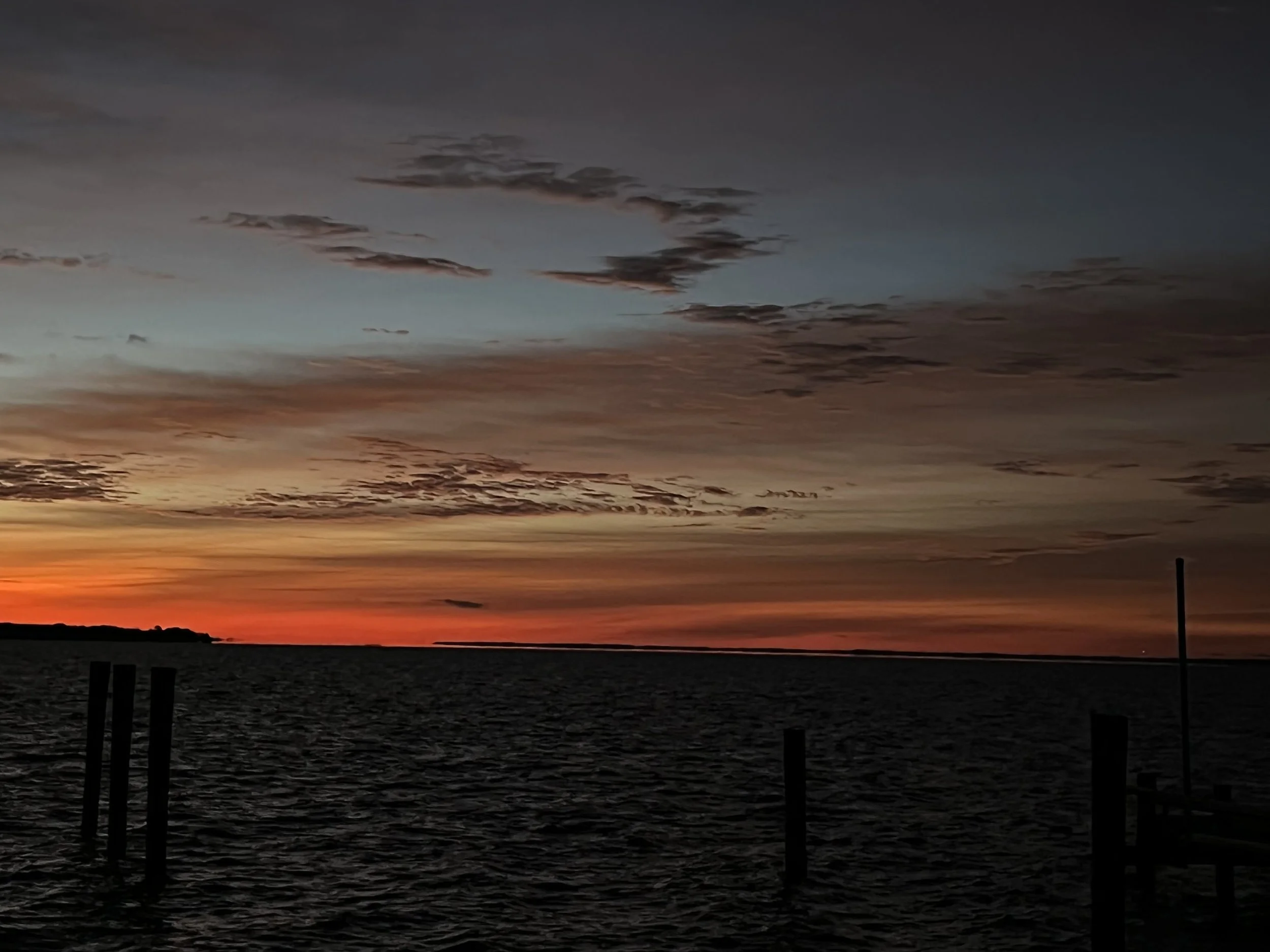 A sunset over the ocean with an orange and red sky, scattered clouds, and a silhouetted shoreline with a wooden dock in the foreground.