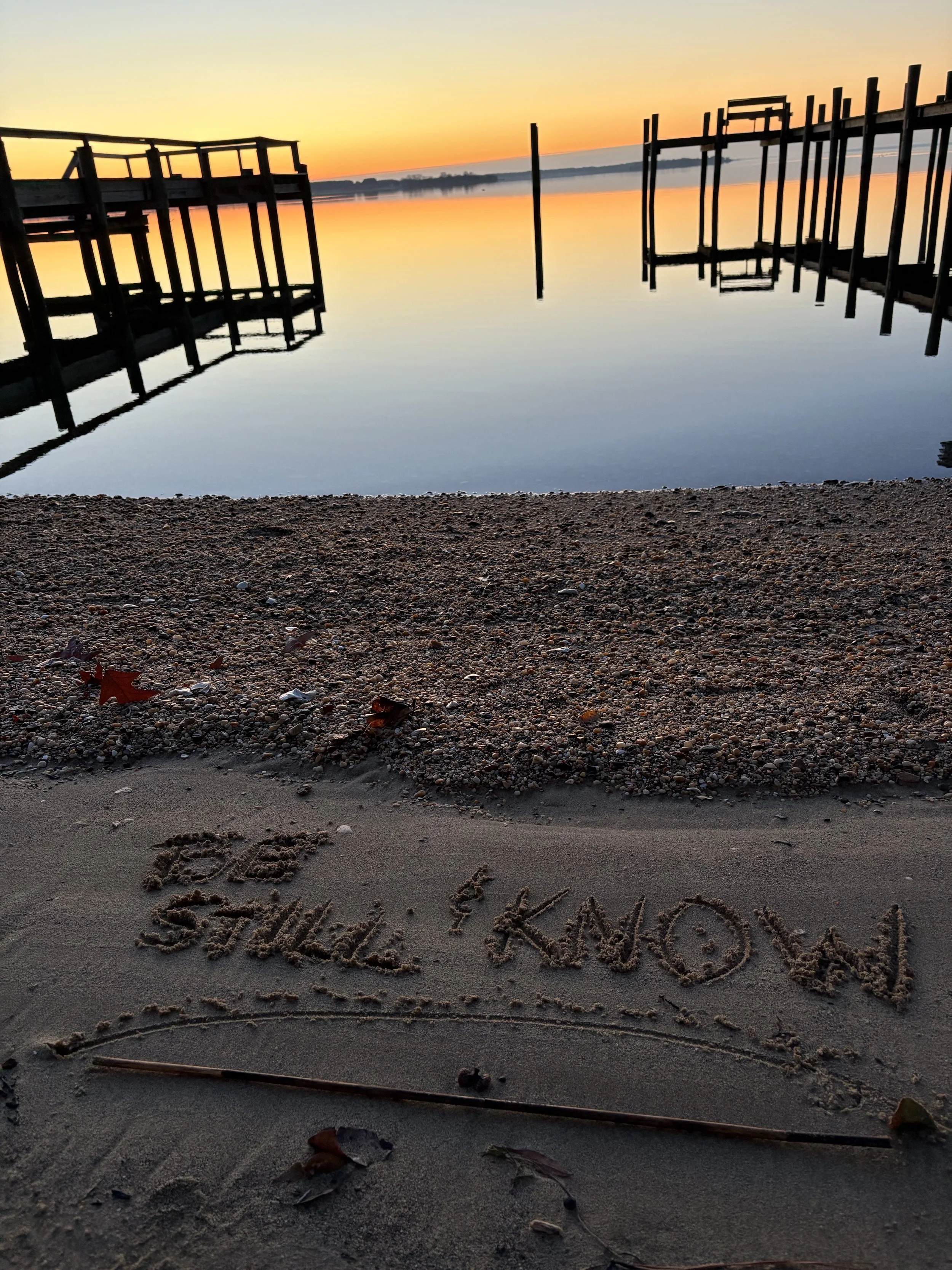 A sunset over a calm lake with a dock on each side reflected in the water. The sky is orange and yellow, and the shoreline has pebbles and fallen leaves, with words written in the sand at the bottom.