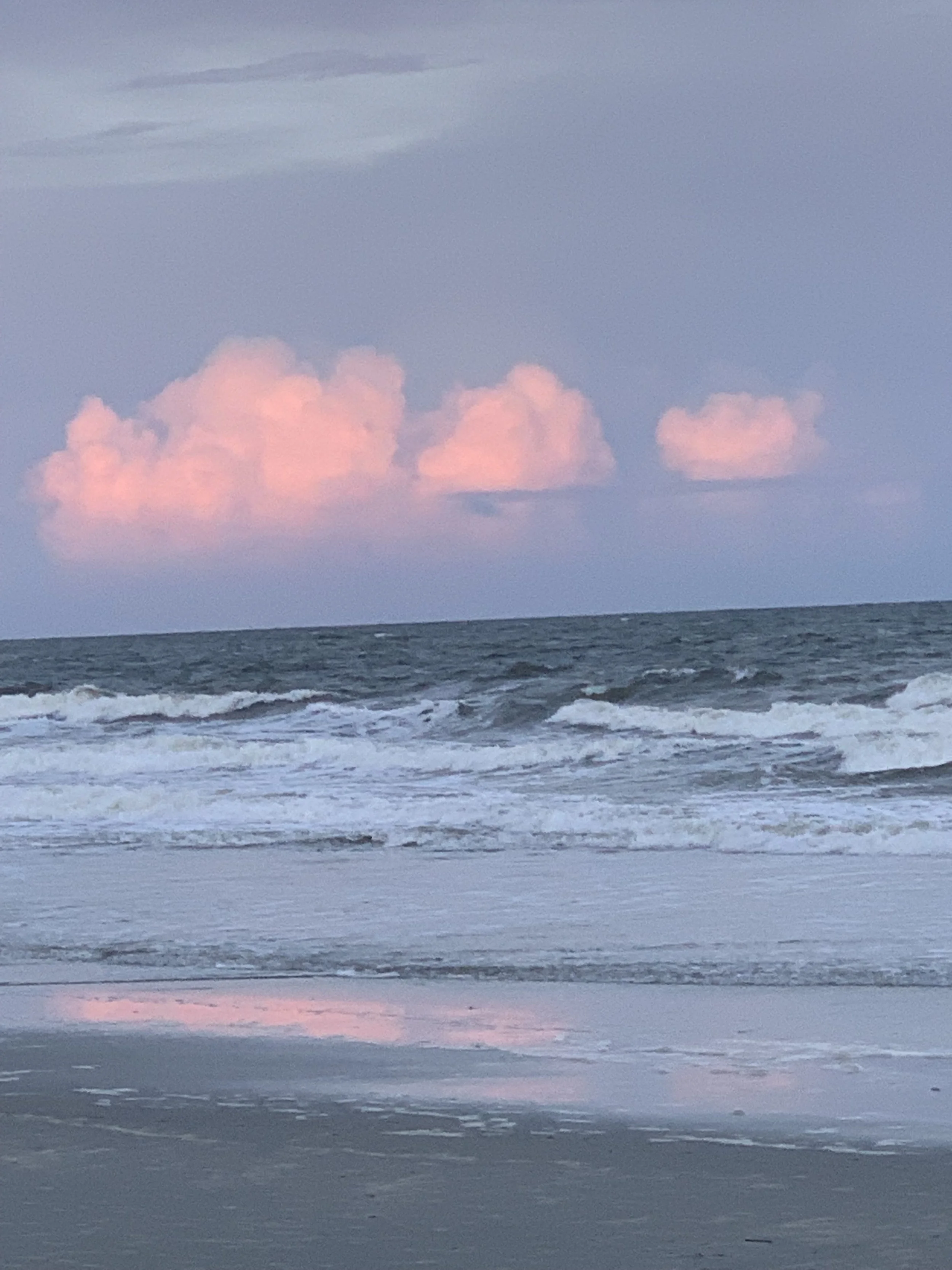A view of the ocean with gentle waves and pink clouds in the sky during dusk or dawn.