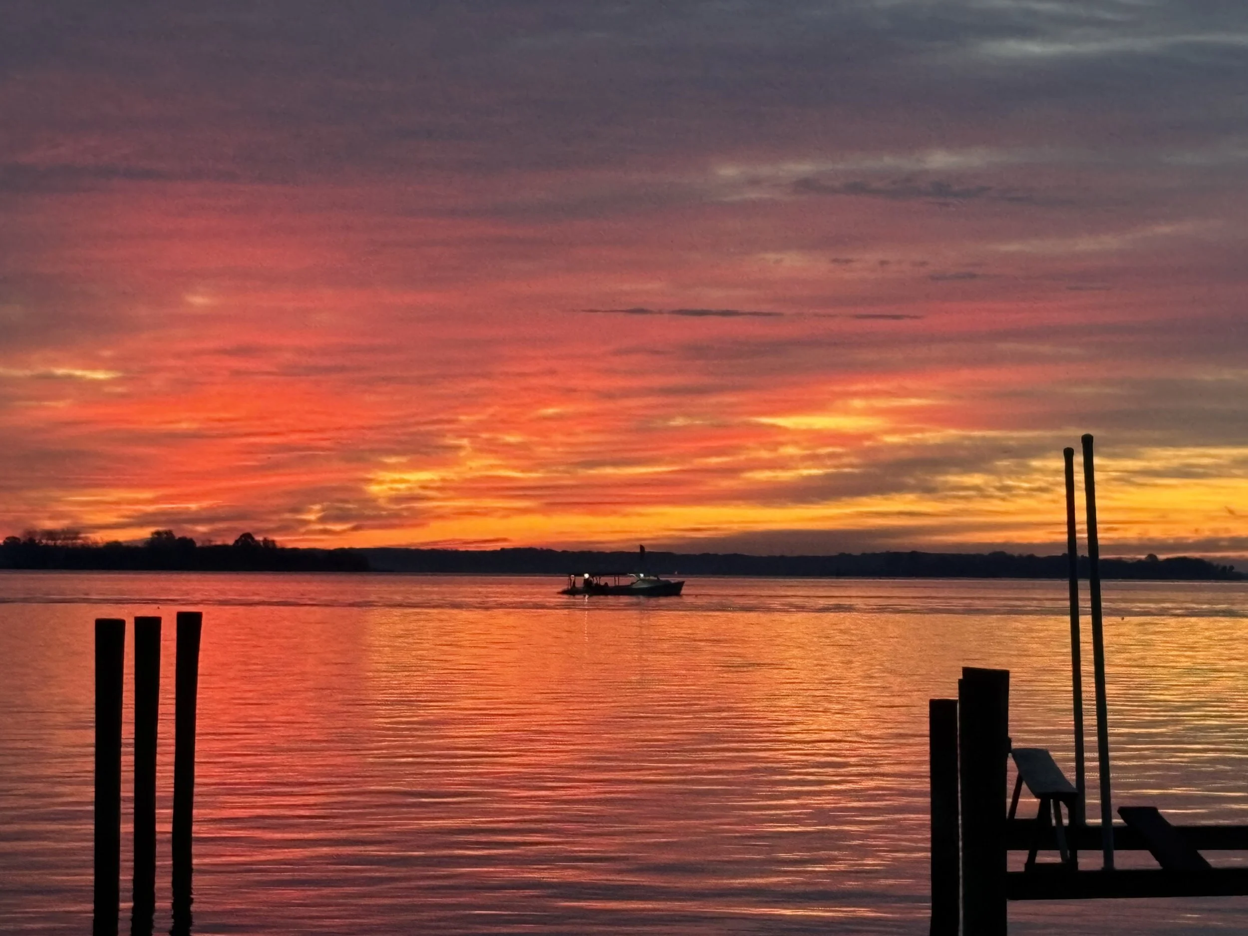 Colorful sunset reflecting over a body of water with two silhouetted poles, a boat with a person, and a chair on the shoreline.