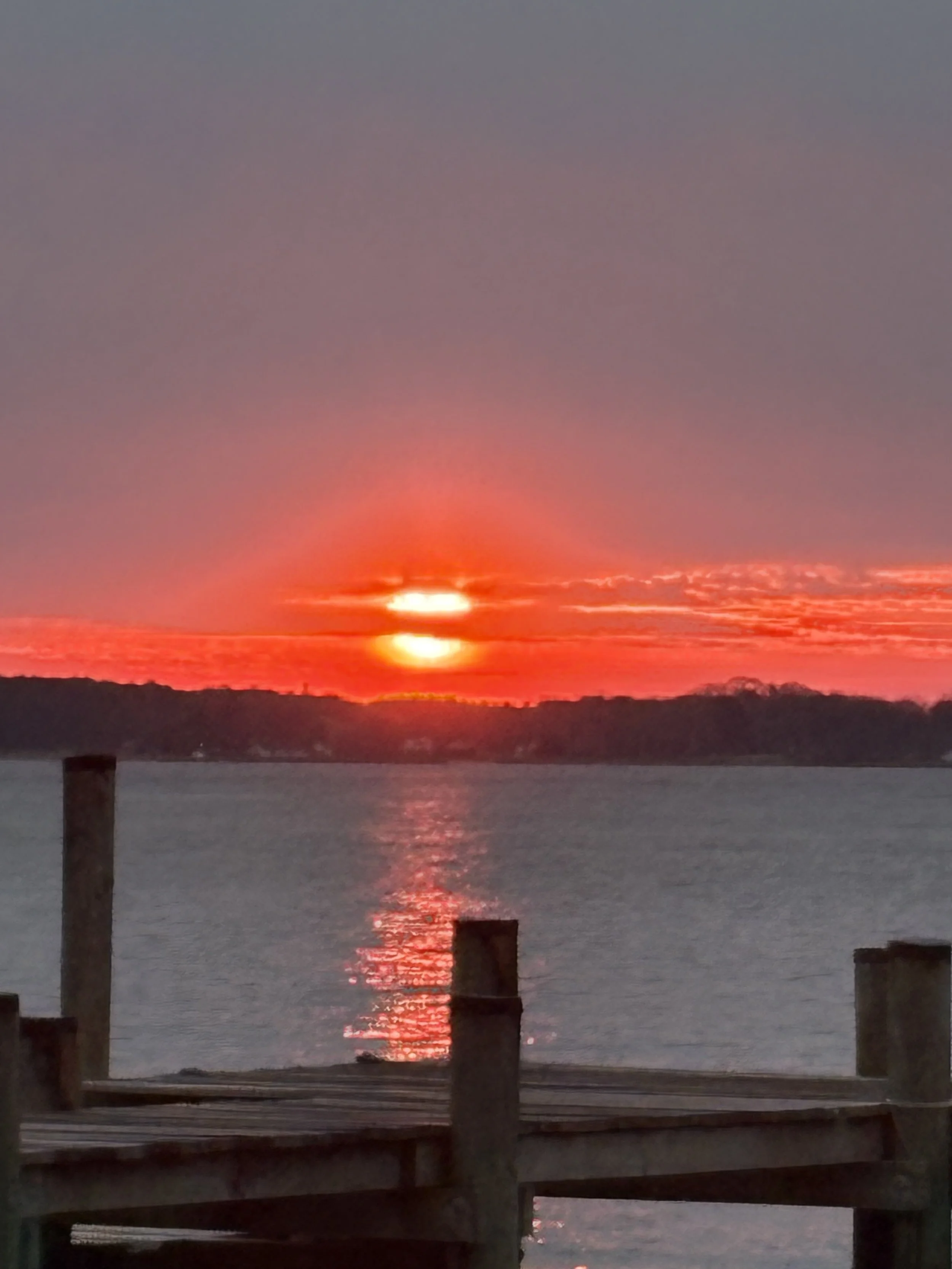 A sunset over a body of water with the sky painted in shades of pink, orange, and purple, and silhouettes of wooden posts and hills in the foreground.