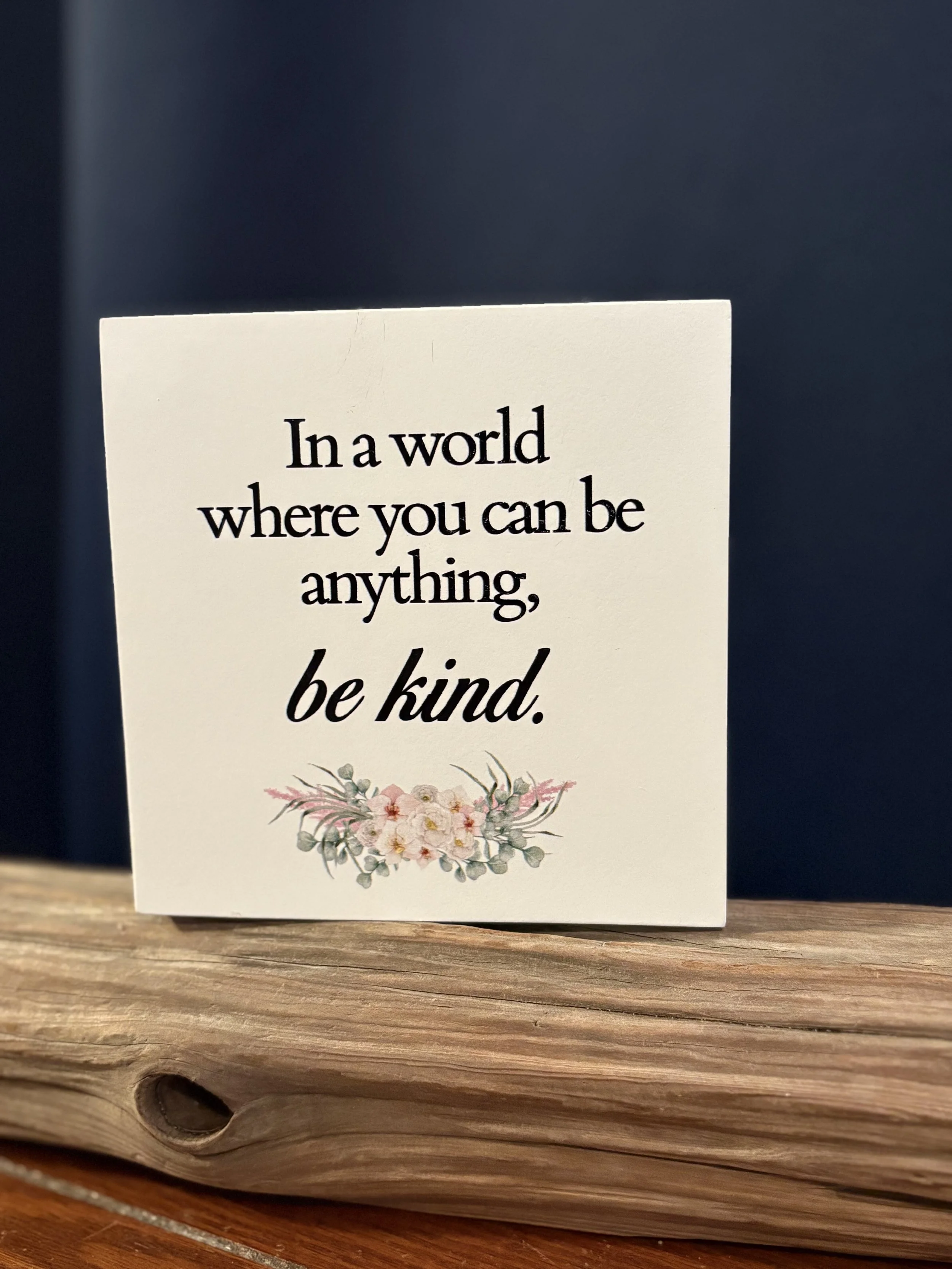 A card with quote text, 'In a world where you can be anything, be kind,' sitting on a rustic wooden surface.
