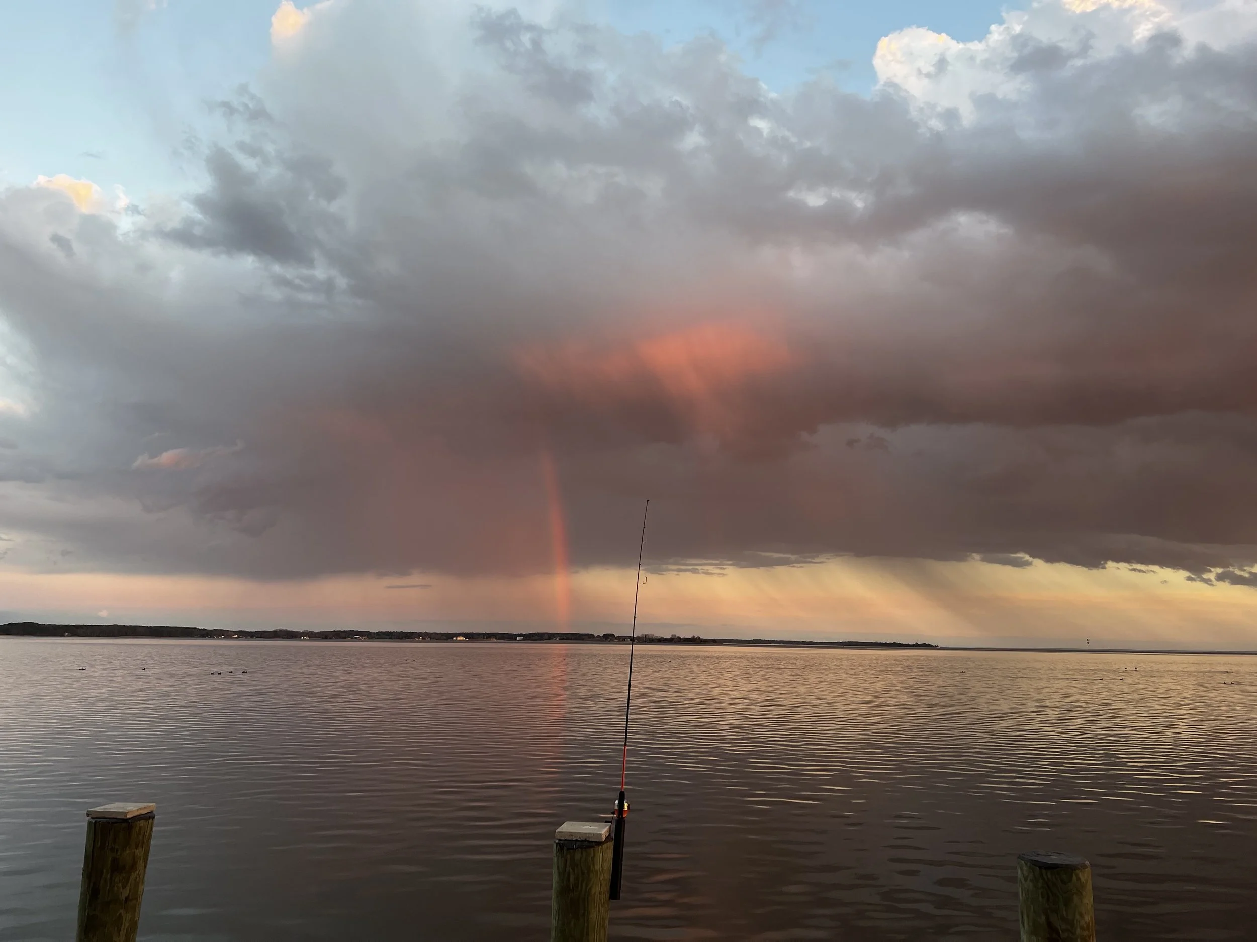 Photo of a body of water with a fishing rod set up on the dock, cloudy sky with patches of blue, and a faint rainbow visible in the sky during sunset or sunrise.