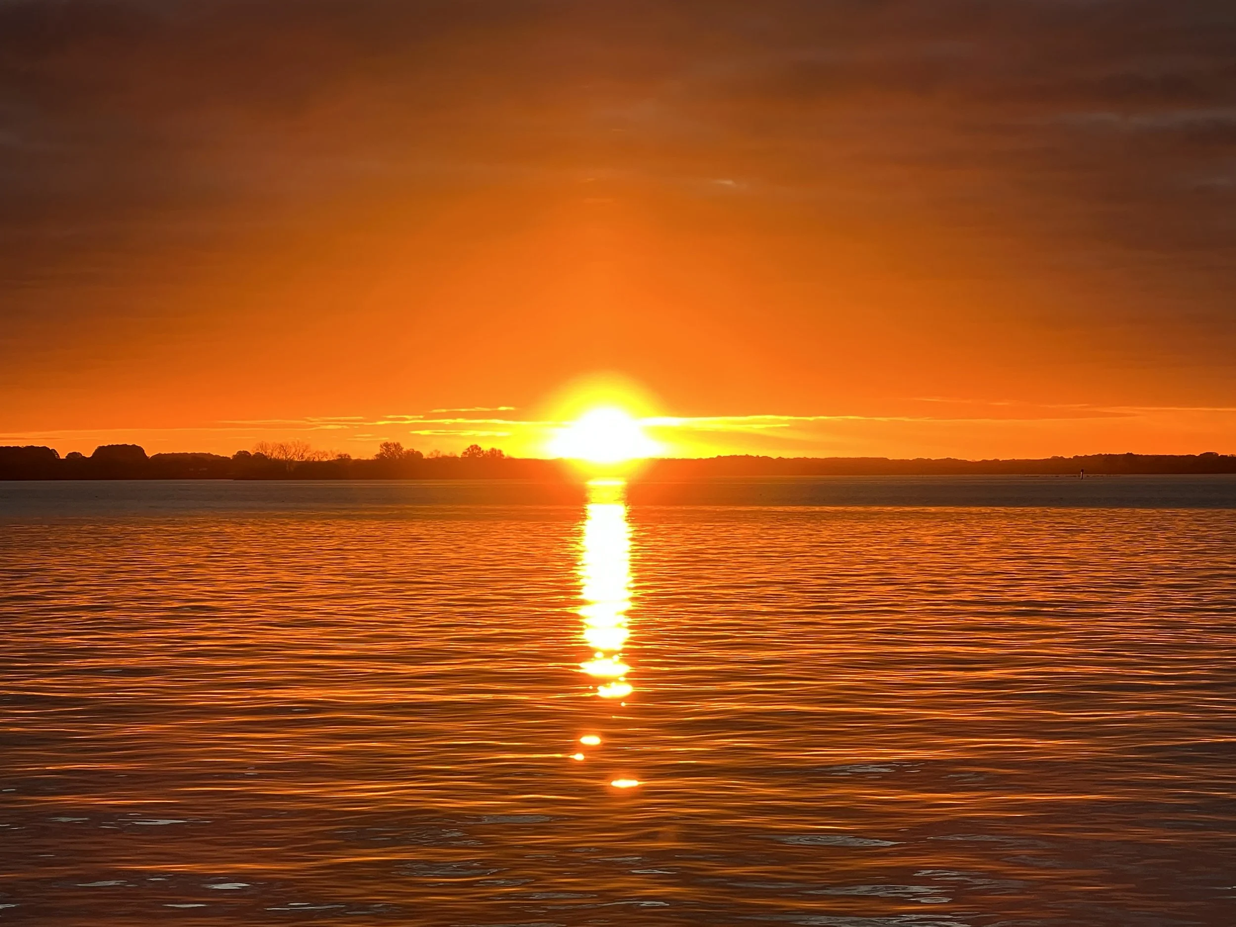 A vibrant sunset over a calm body of water with trees on the horizon.