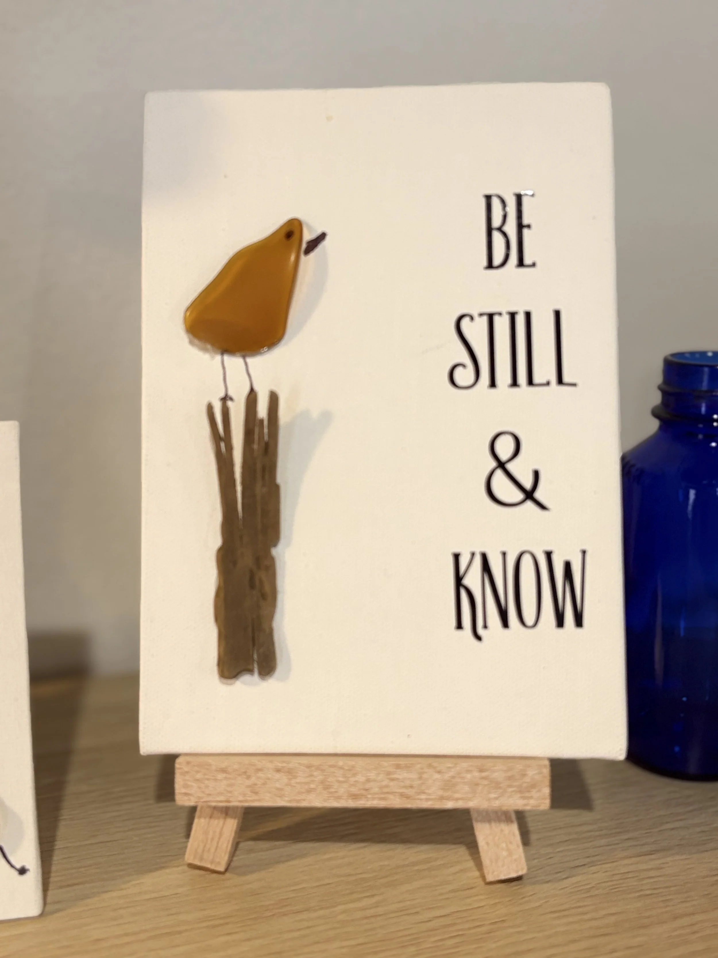 Wooden stand holding a white sign with black letters that reads "BE STILL & KNOW." An almond-shaped yellow decorative object with a small black eye and beak, resembling a bird, is attached to the sign. Part of a blue bottle is visible on the right.