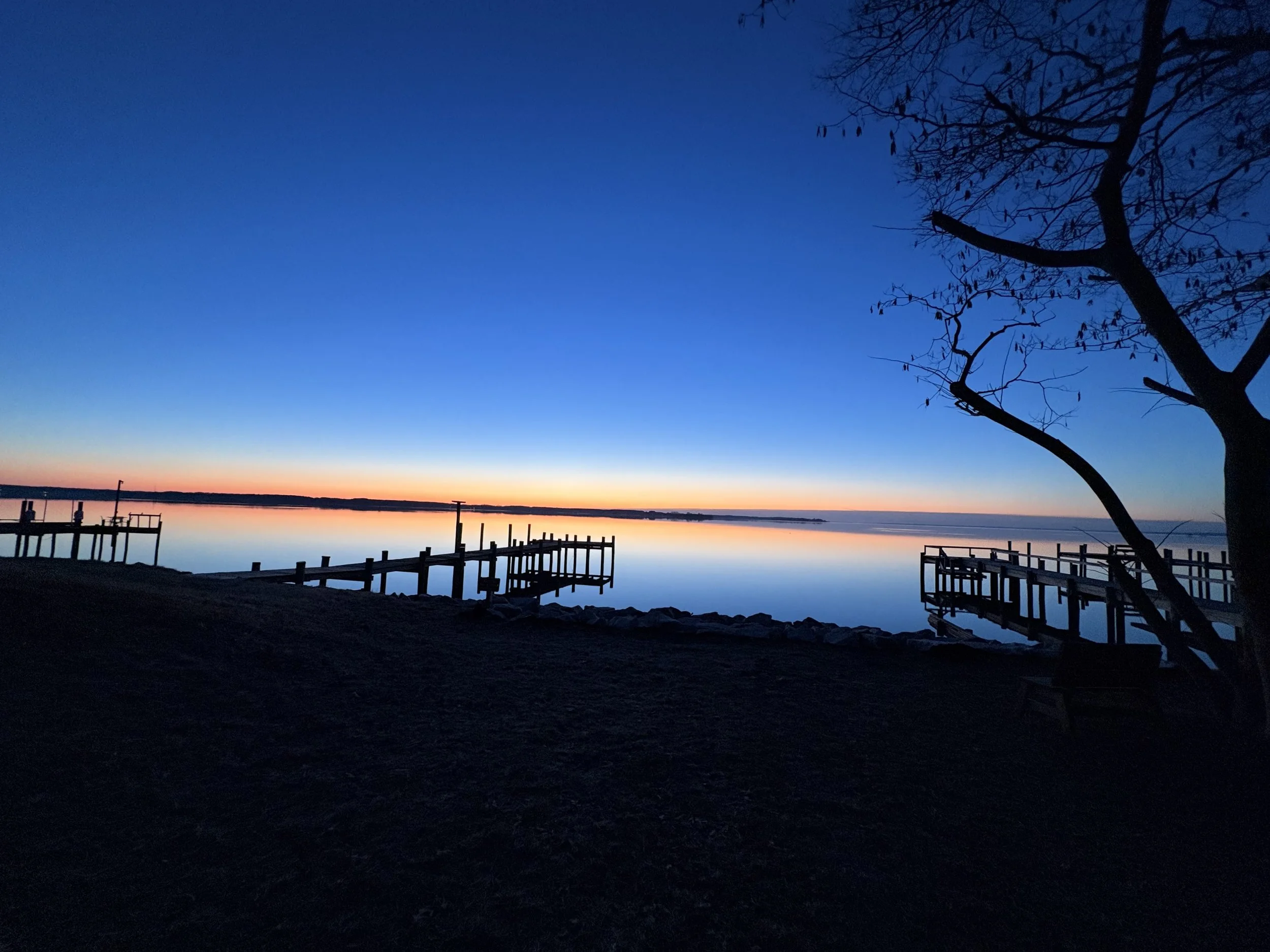 A peaceful lakeside scene at sunset with a silhouette of a tree on the right, docks extending into still water, and a colorful orange and pink horizon in the background.