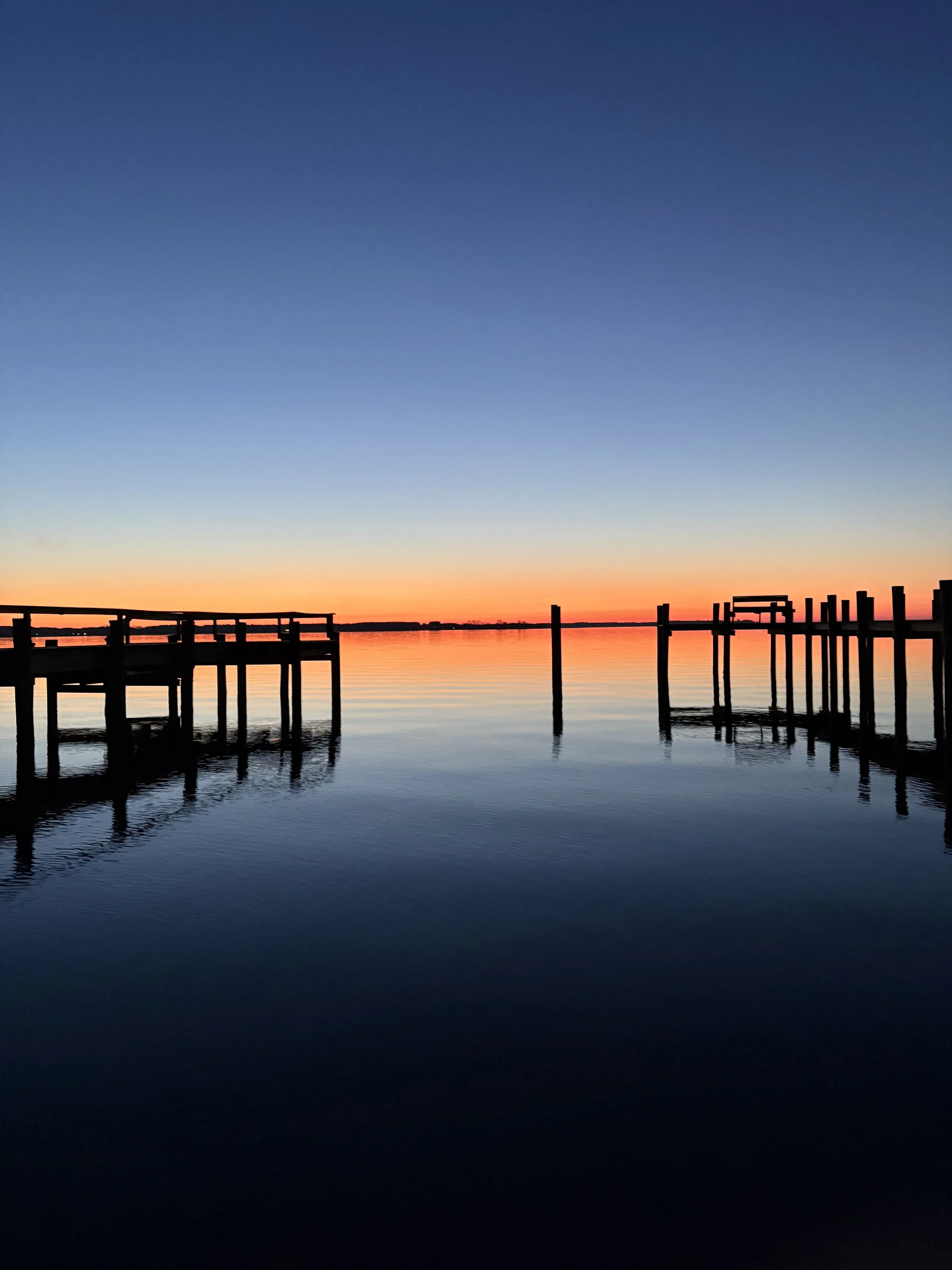 Silhouettes of wooden dock structures stretching into calm water during sunrise with a colorful sky fading from orange to deep blue.