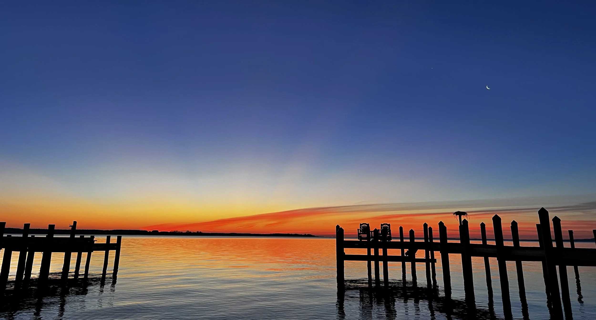 A sunset over calm water with a colorful sky transitioning from orange to blue, with silhouetted wooden docks and chairs in the foreground.