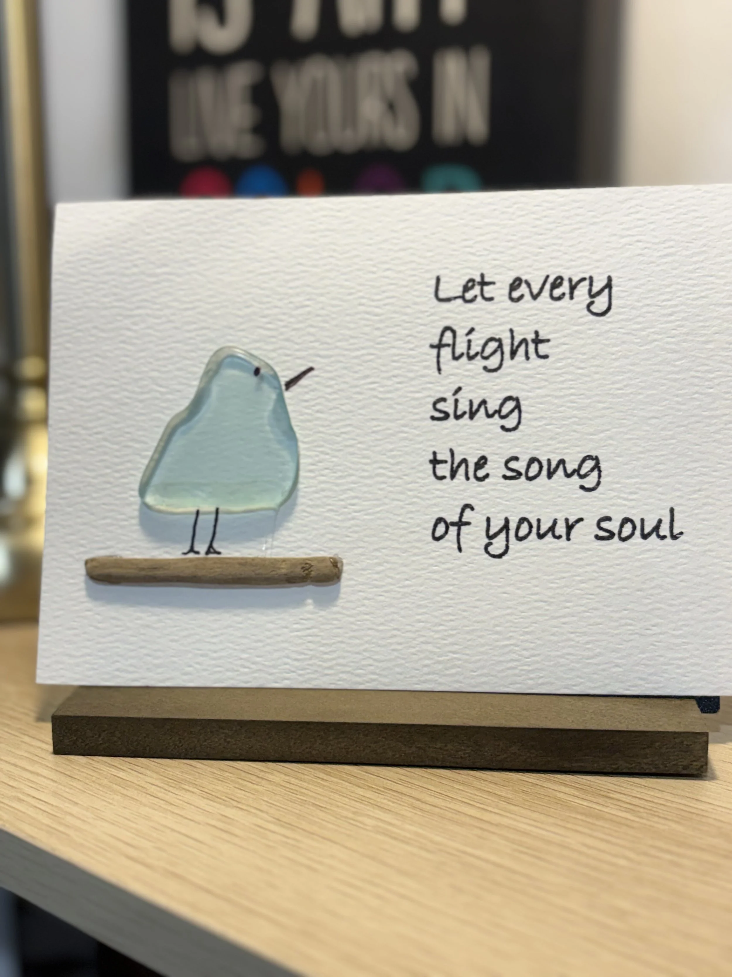 A greeting card on a wooden surface features an illustration of a light blue bird with a small beak, a black eye, and a tiny black line for a wing. Next to the bird, handwritten-style text reads: "Let every flight sing the song of your soul."