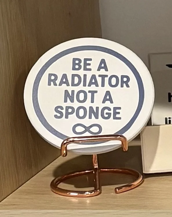A circular sign on a wooden surface that says "Be a radiator not a sponge" with an infinity symbol below the text.