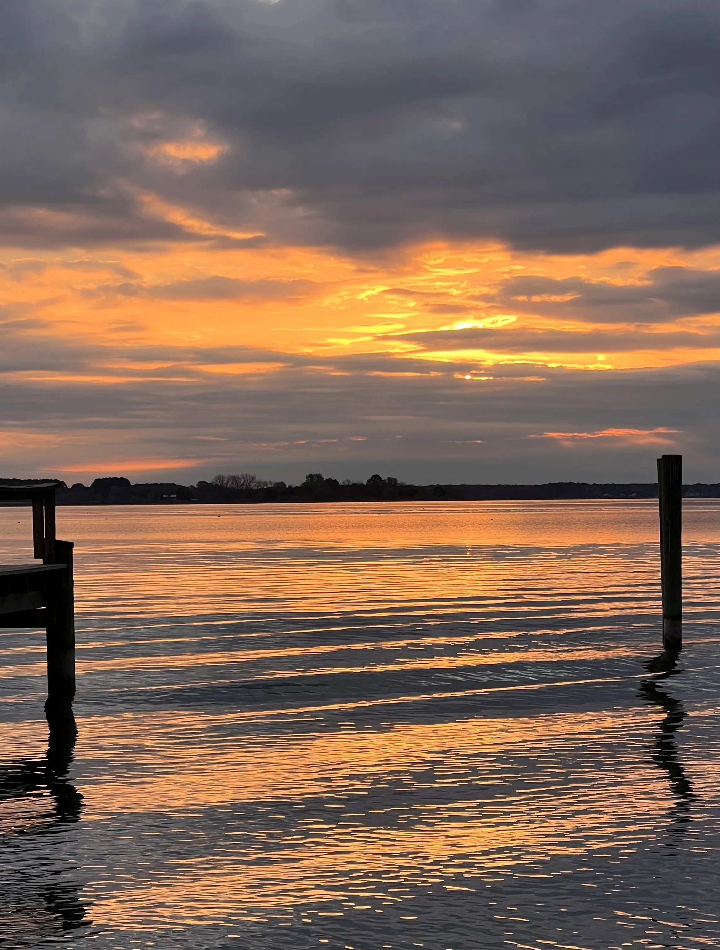 A sunset over a body of water, with dark clouds overhead and orange and yellow hues reflecting on the water surface, framed by wooden posts at the dock.
