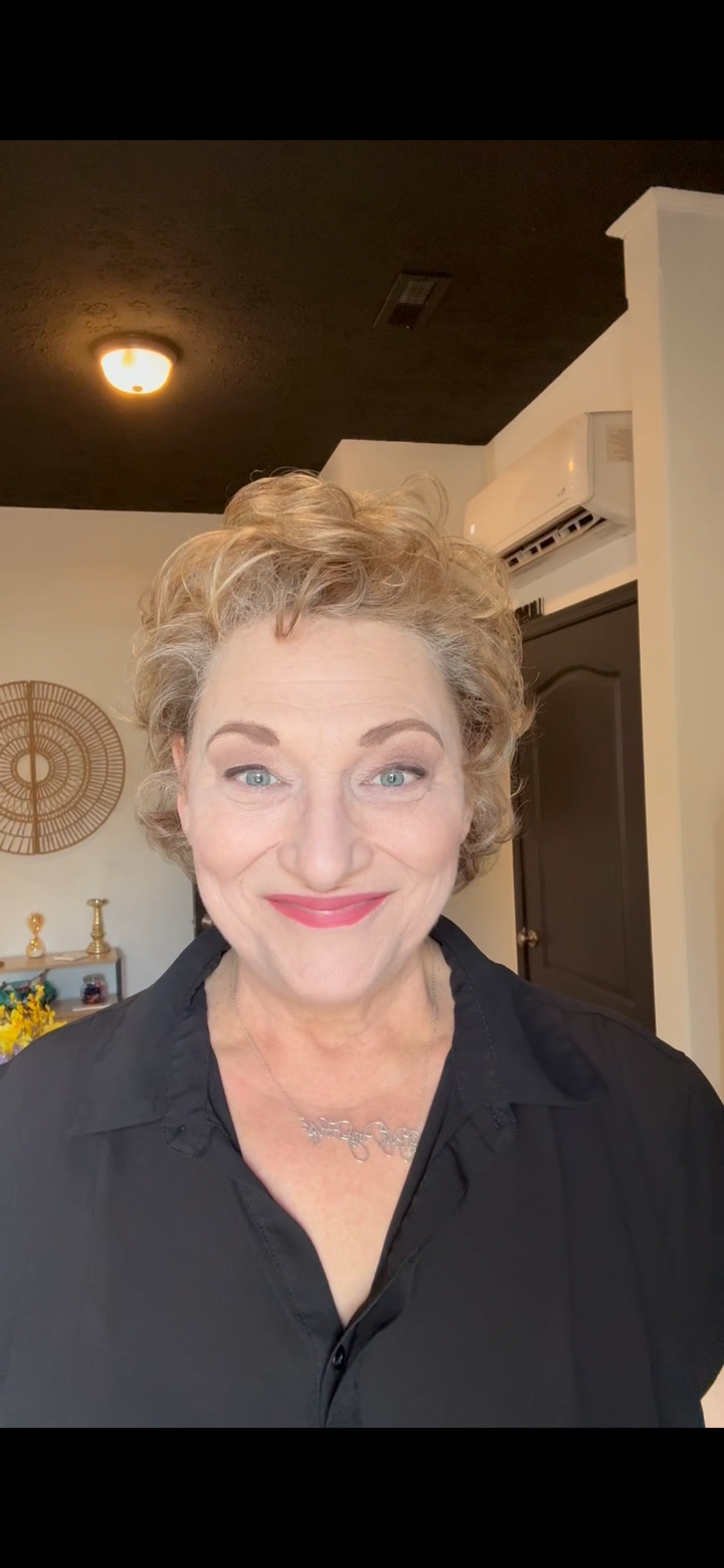 Portrait of a middle-aged woman with short curly blonde hair and blue eyes, smiling and wearing a black top and a silver necklace, in a cozy interior setting.