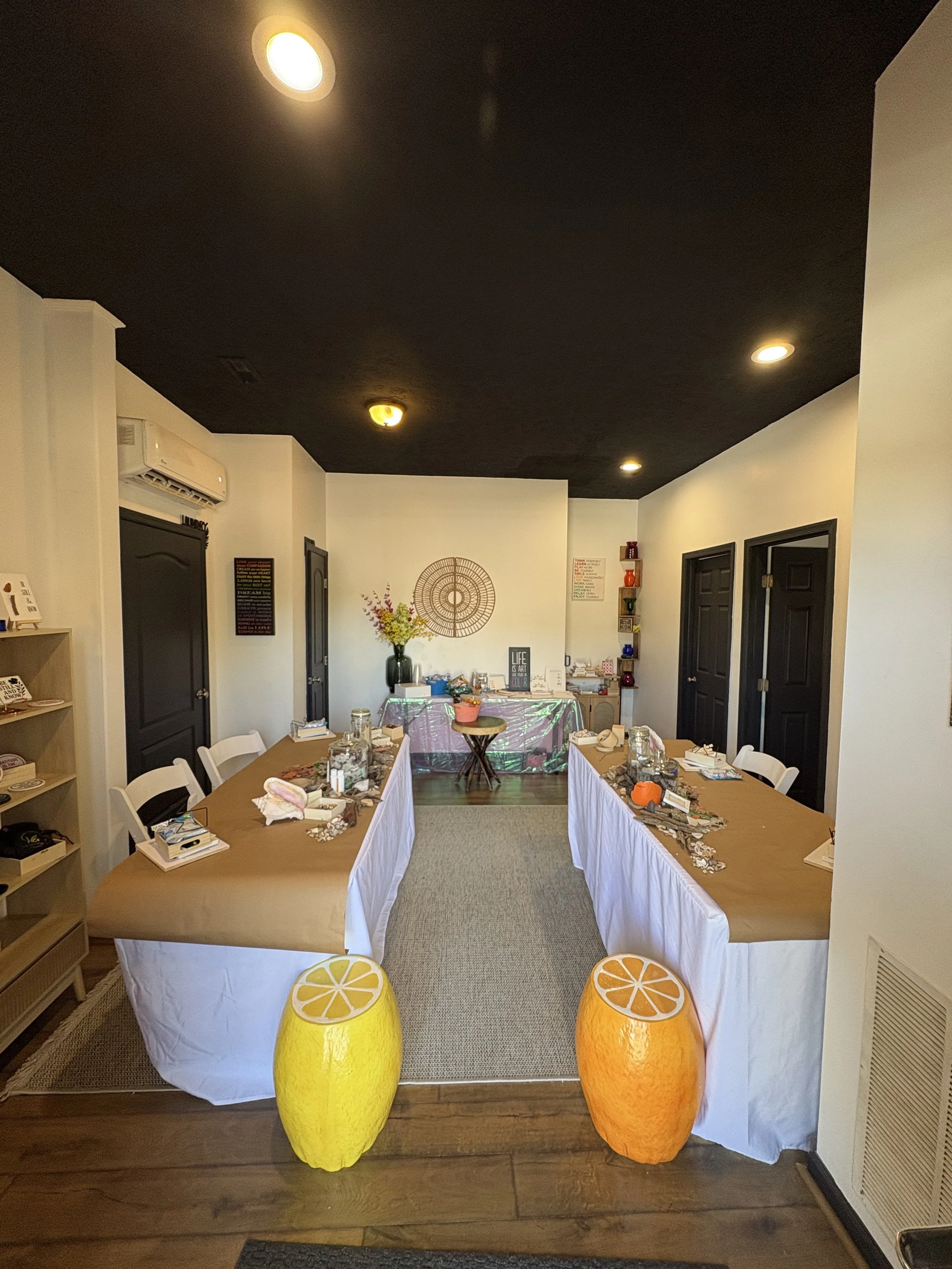 A room decorated for a gathering, with two tables draped in white tablecloths, set with mason jars, small decorations, and books. In front of the tables are two large, decorative lemon and orange-shaped stools. The back wall features a flower arrange