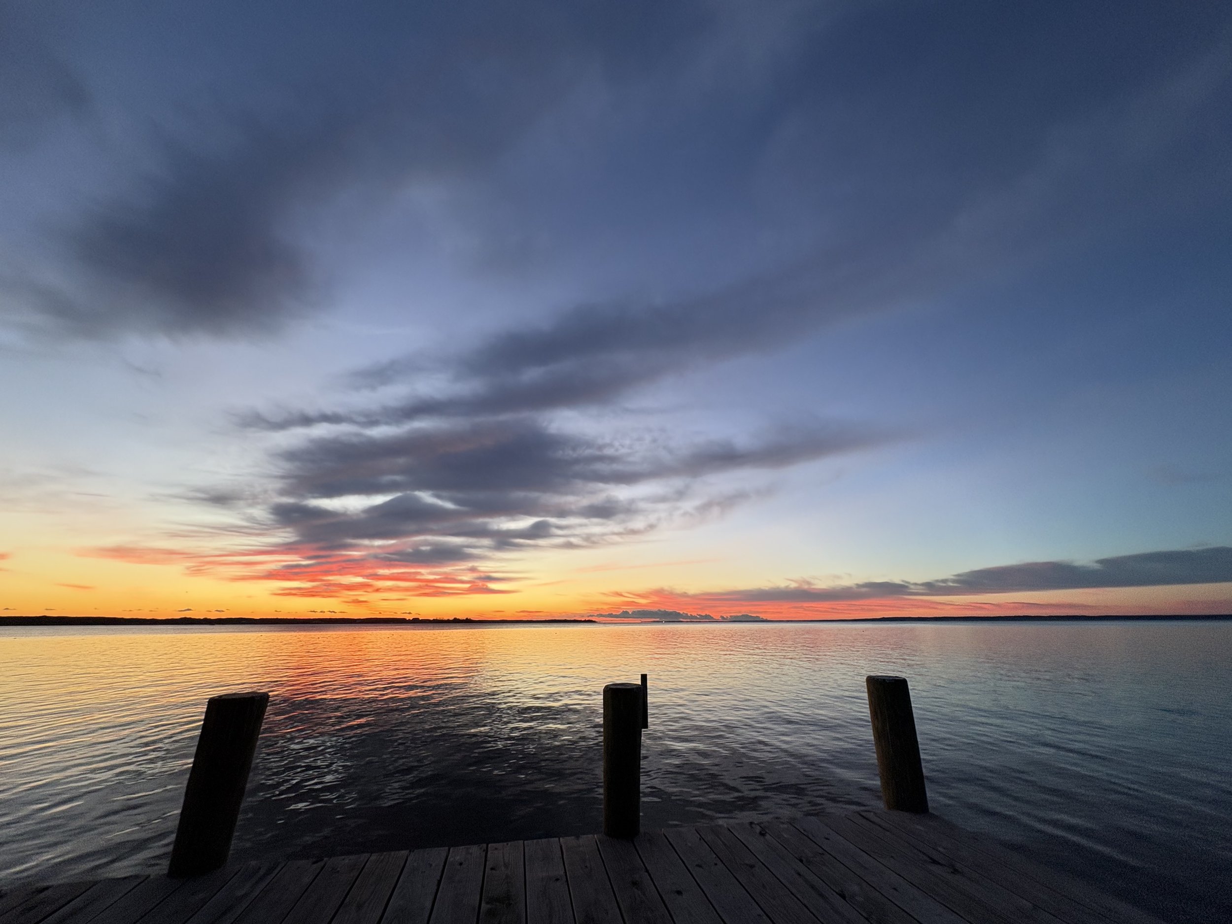 A scenic view of a sunset over a body of water, with dark clouds in the sky and a wooden dock in the foreground.