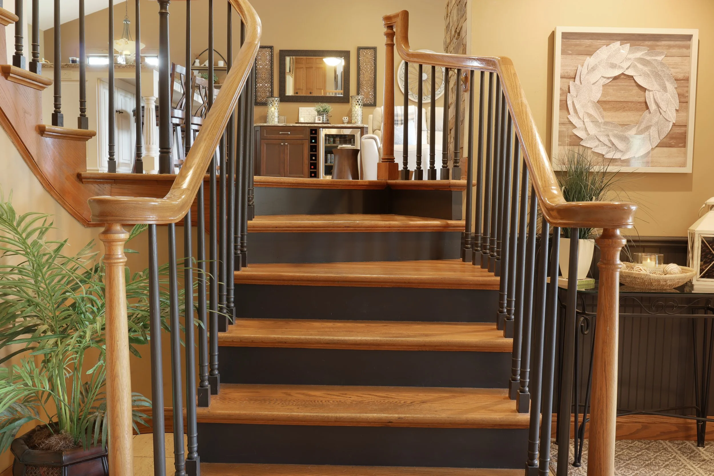 Interior view of a staircase with wooden steps and black railings, leading to a living space with a kitchen and dining area. The background includes wall art, a mirror, and plants.