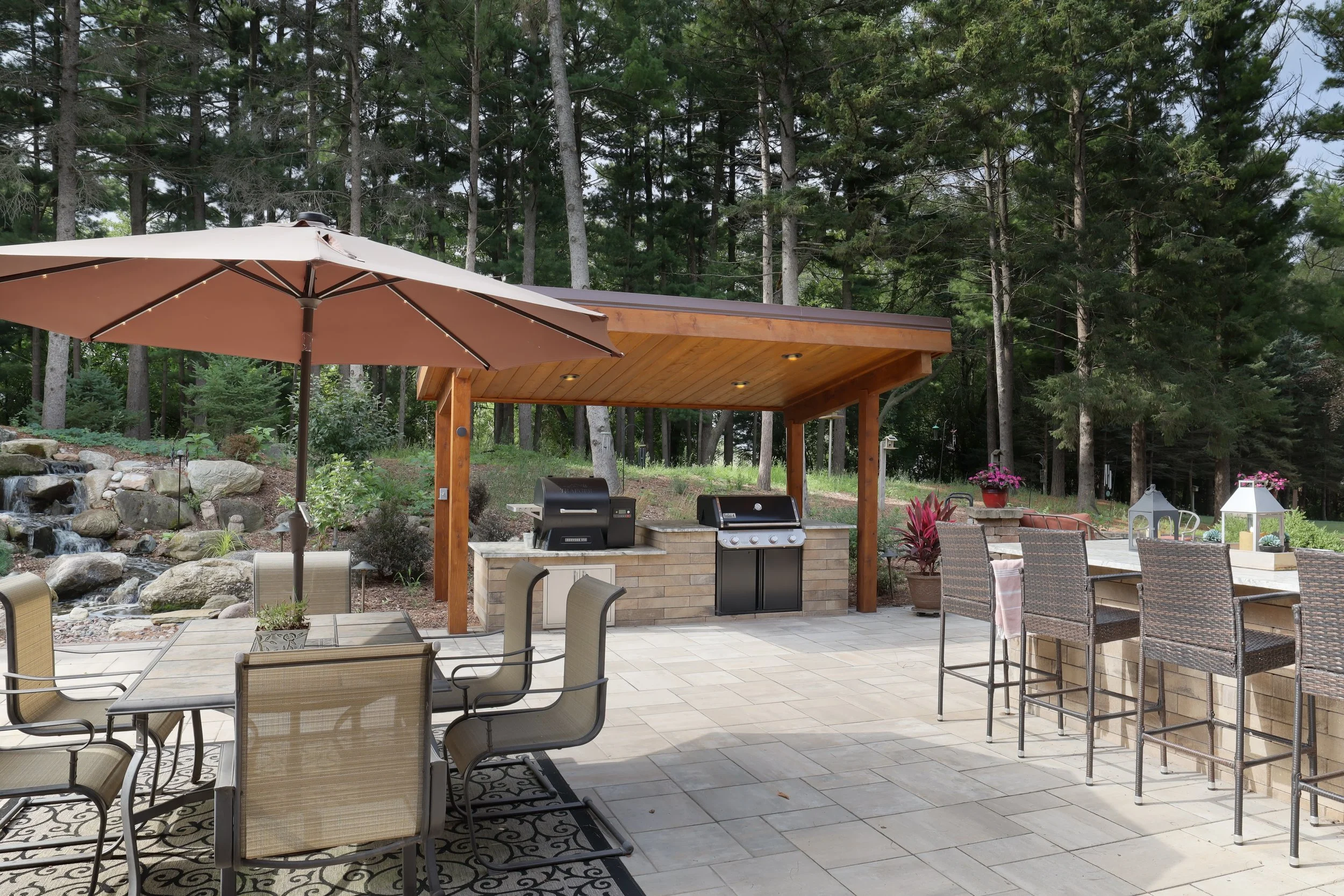 An outdoor patio area with a covered wooden pergola, a grill, and various chairs and tables. There is a large beige umbrella and a garden scene with trees and flowers in the background.