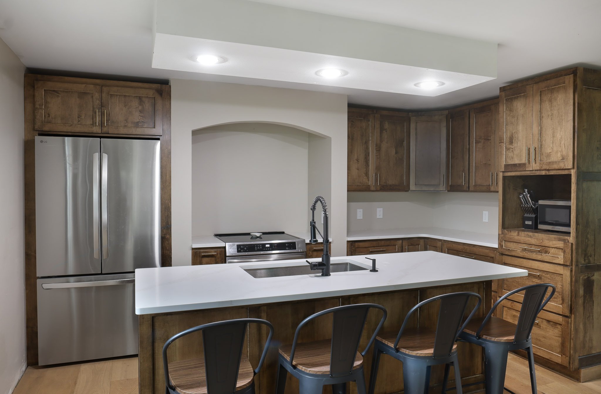 A modern kitchen with white countertops, wooden cabinets, a stainless steel refrigerator, a built-in microwave, and barstools at the counter.