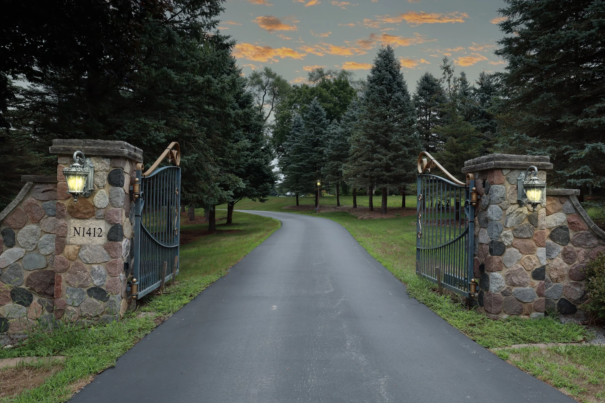 A stone gate with black iron gates opening onto a paved driveway lined with green grass and dense evergreen trees, with a sunset sky with orange clouds in the background.