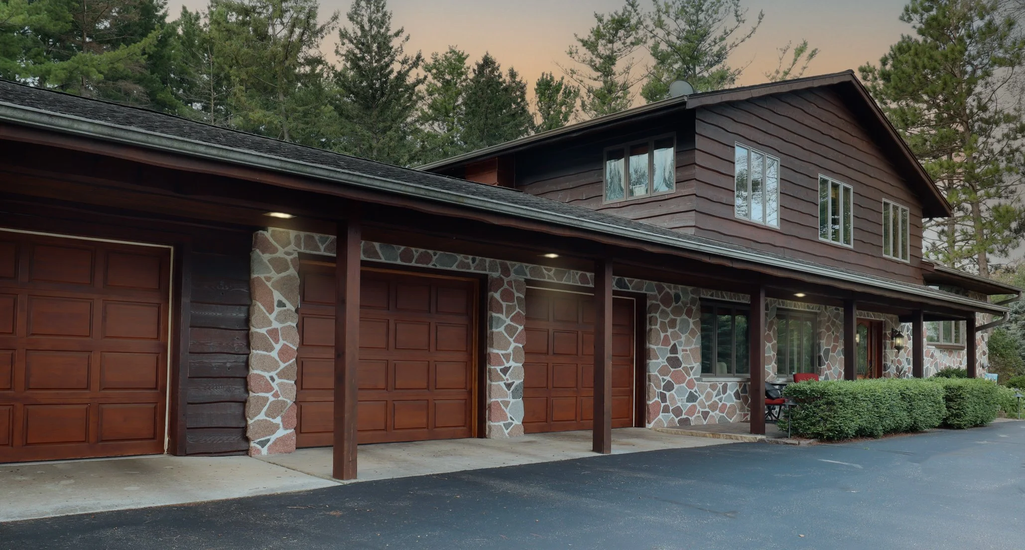 A two-story house with stone and wood exterior, featuring a three-car garage, surrounded by greenery and tall trees, with a driveway in front and a sitting area on the right.