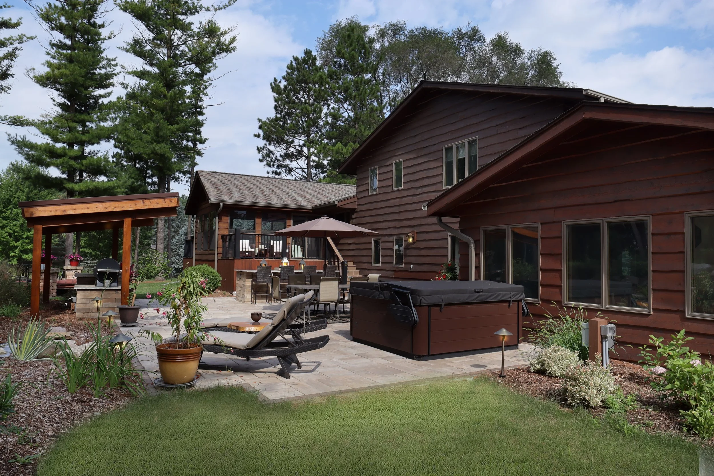 A backyard patio area with outdoor furniture, including a table with chairs, an umbrella, and a hot tub, surrounded by a garden with potted plants and flowers, with a house and tall trees in the background.