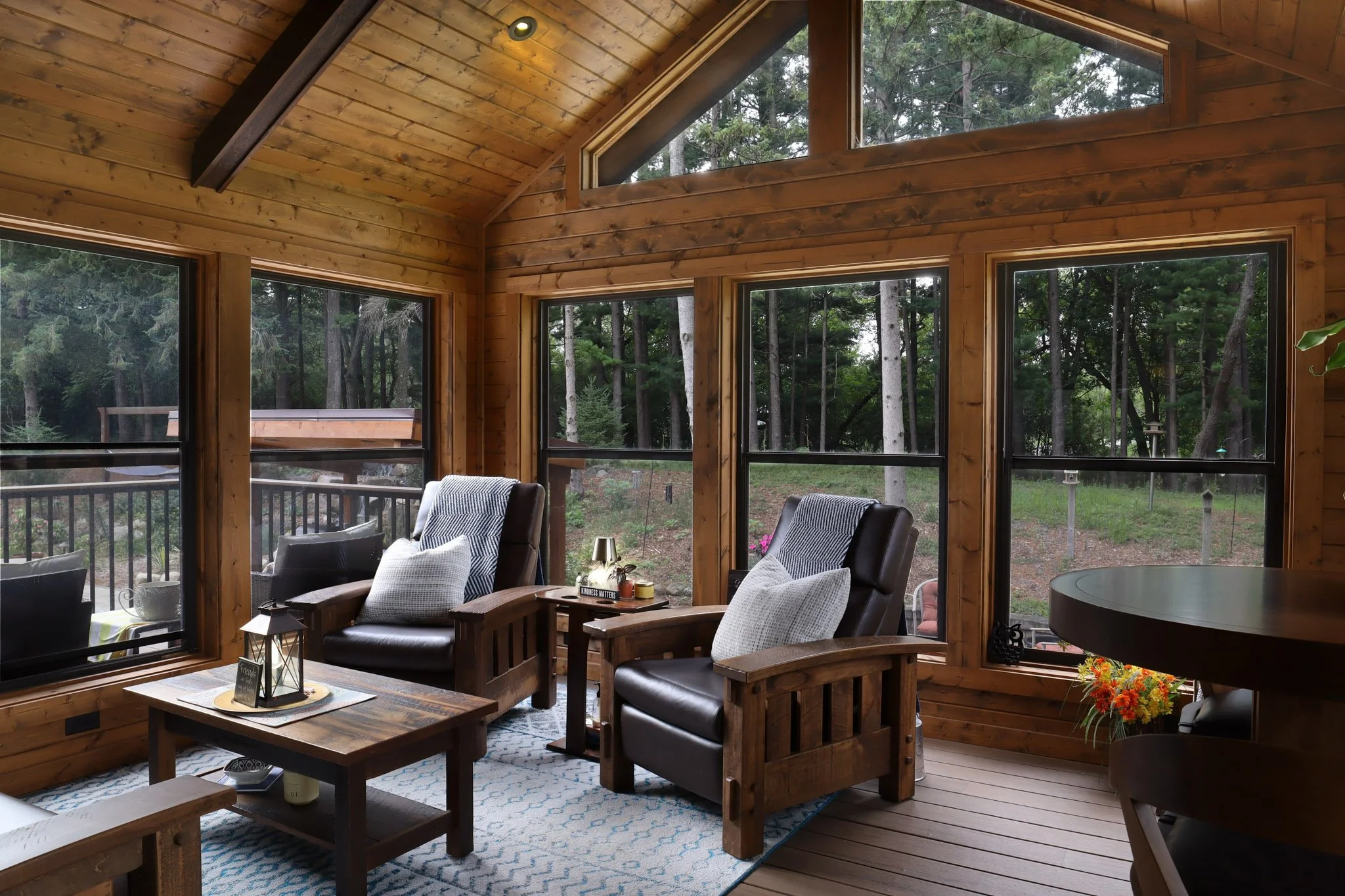 Interior of a wooden house with large windows showing a forest outside, furnished with chairs, a coffee table, and a bright, cozy atmosphere.