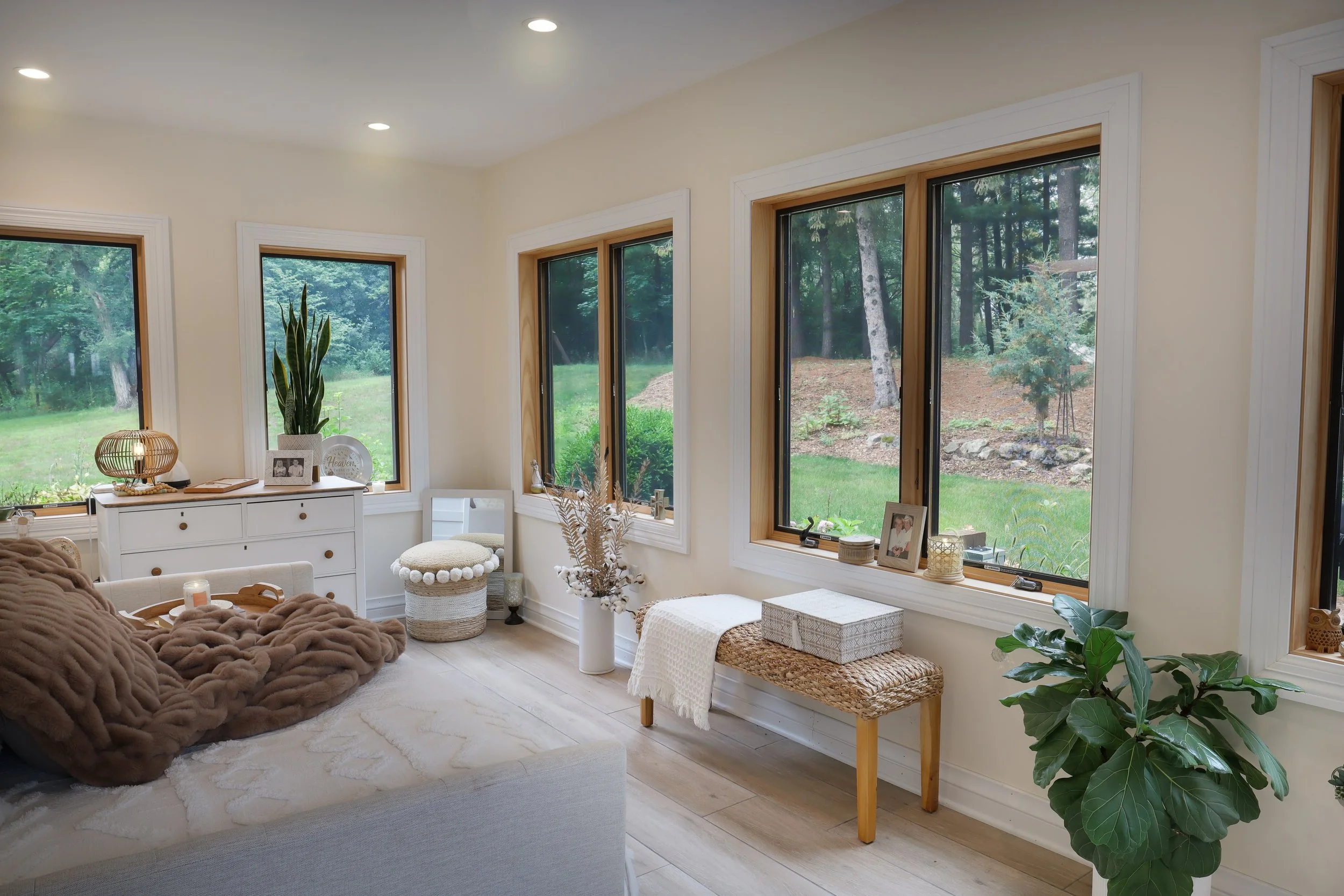 Living room with large windows showing outdoor greenery, beige walls, white furniture, decorative plants, and cozy throws.