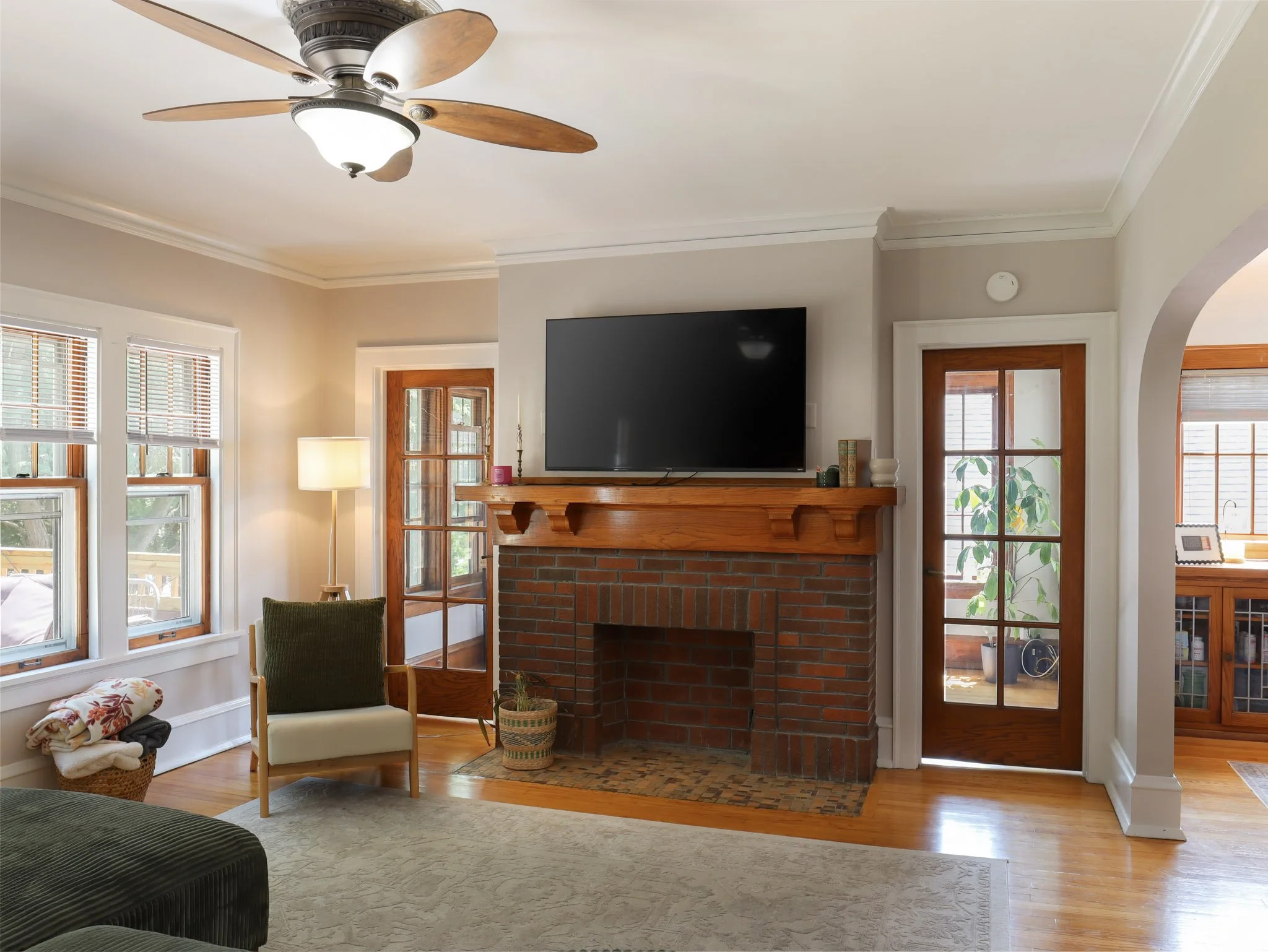 Living room with a ceiling fan, large windows with blinds, a brick fireplace with a mounted flat-screen TV, a green armchair, a cream chair, a lamp, a basket of blankets, and wooden floors.