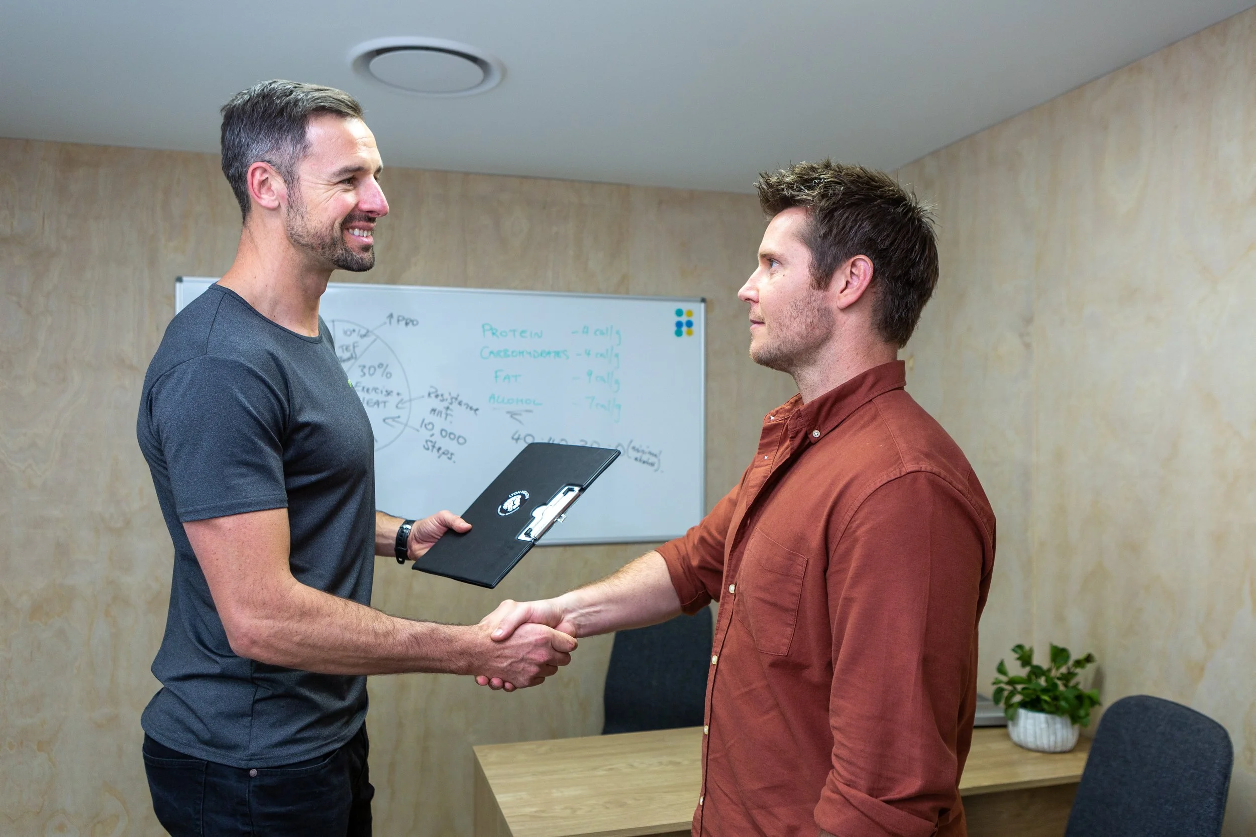 Two men shaking hands in a meeting room, with one handing the other a black folder. In the background, there is a whiteboard with notes and a small potted plant on a table.