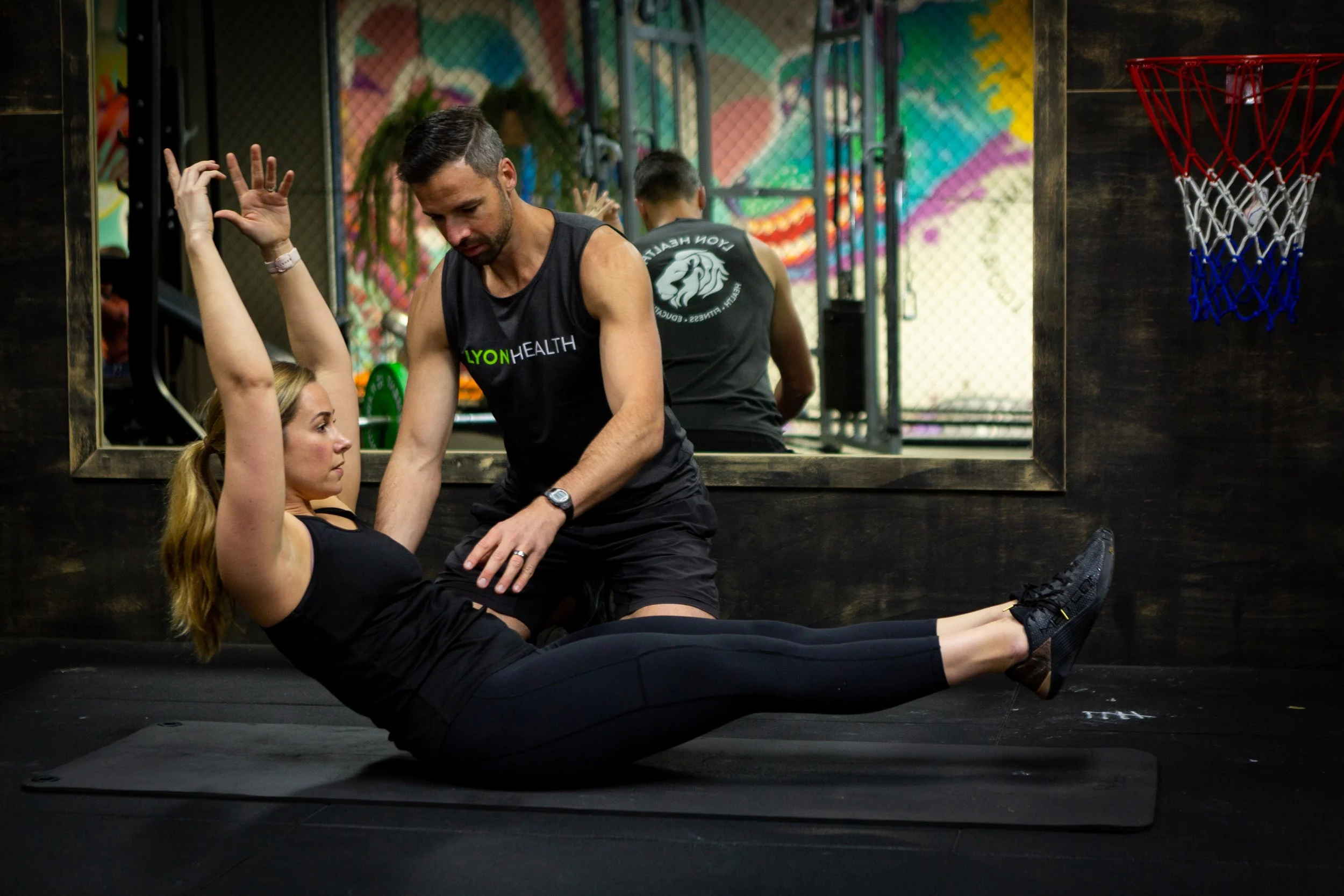 A man and woman exercising in a gym. The woman is sitting on the floor, stretching her arm, while the man is leaning over her, holding her arm. There is a large mirror behind them, reflecting colorful graffiti on the wall and a basketball hoop.
