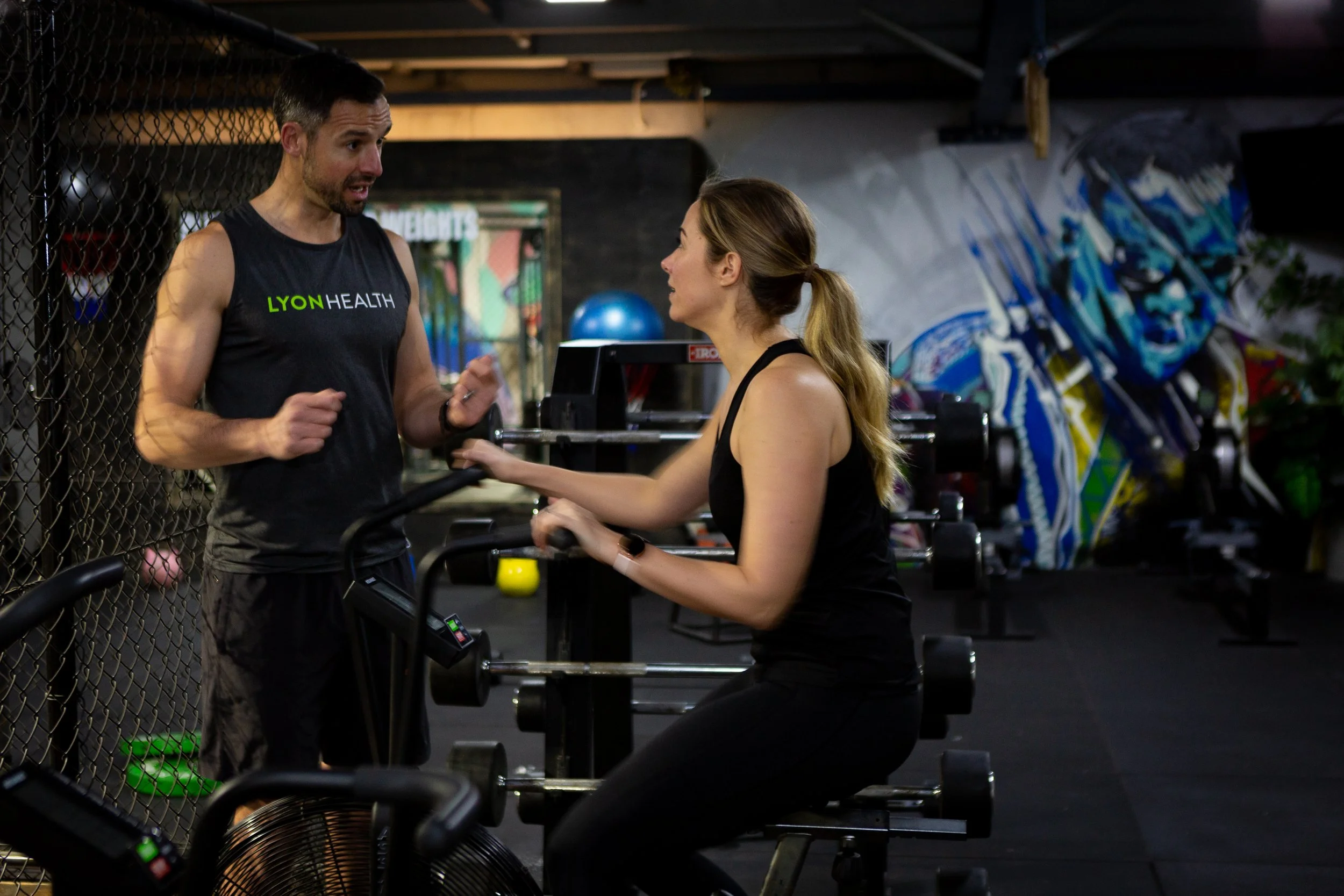 A man and woman are having a conversation in a gym. The man is wearing a black sleeveless shirt with