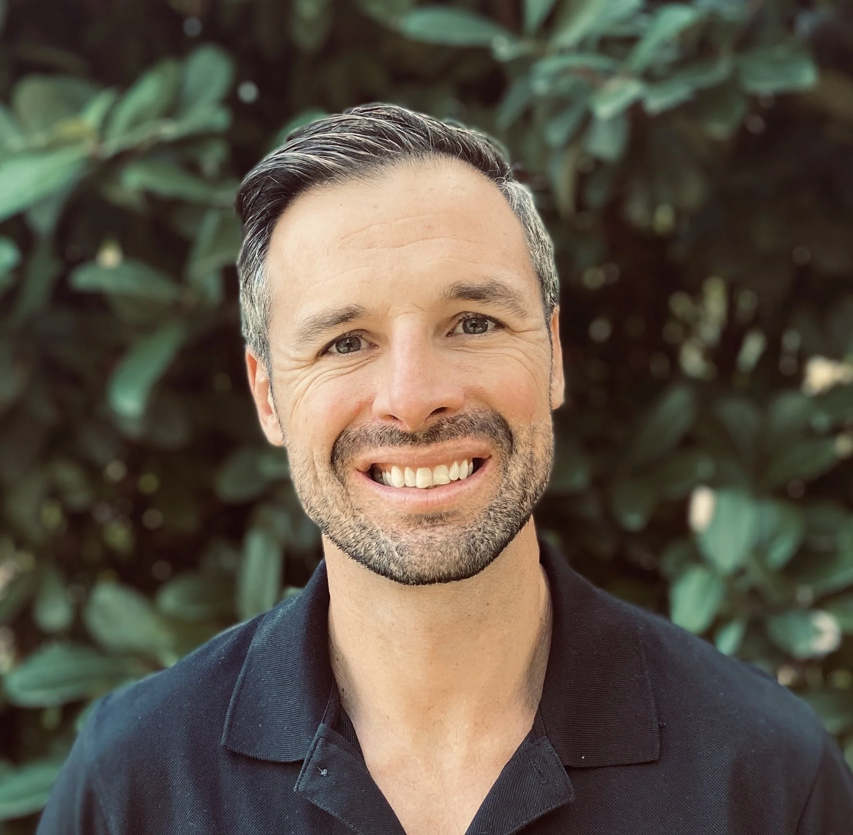 A smiling man with short black hair, gray streaks, and facial hair, wearing a black shirt, standing outdoors with green foliage background.