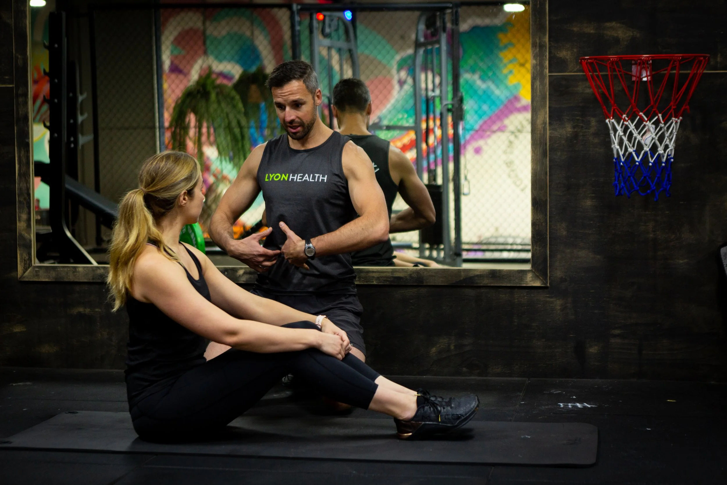 A man and a woman are in a gym, having a conversation. The woman is seated on the floor doing a stretching exercise, and the man stands near her. The background features a mirror, colorful graffiti, and a basketball hoop on the wall.