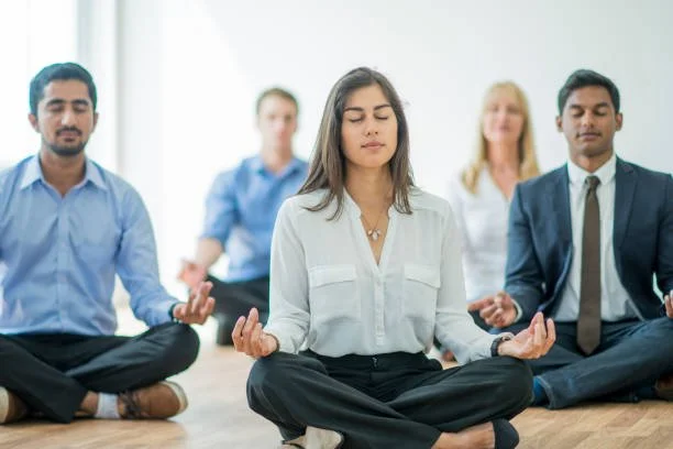 Group of five diverse adults practicing meditation or yoga in a bright room, sitting cross-legged with eyes closed and hands resting on knees.