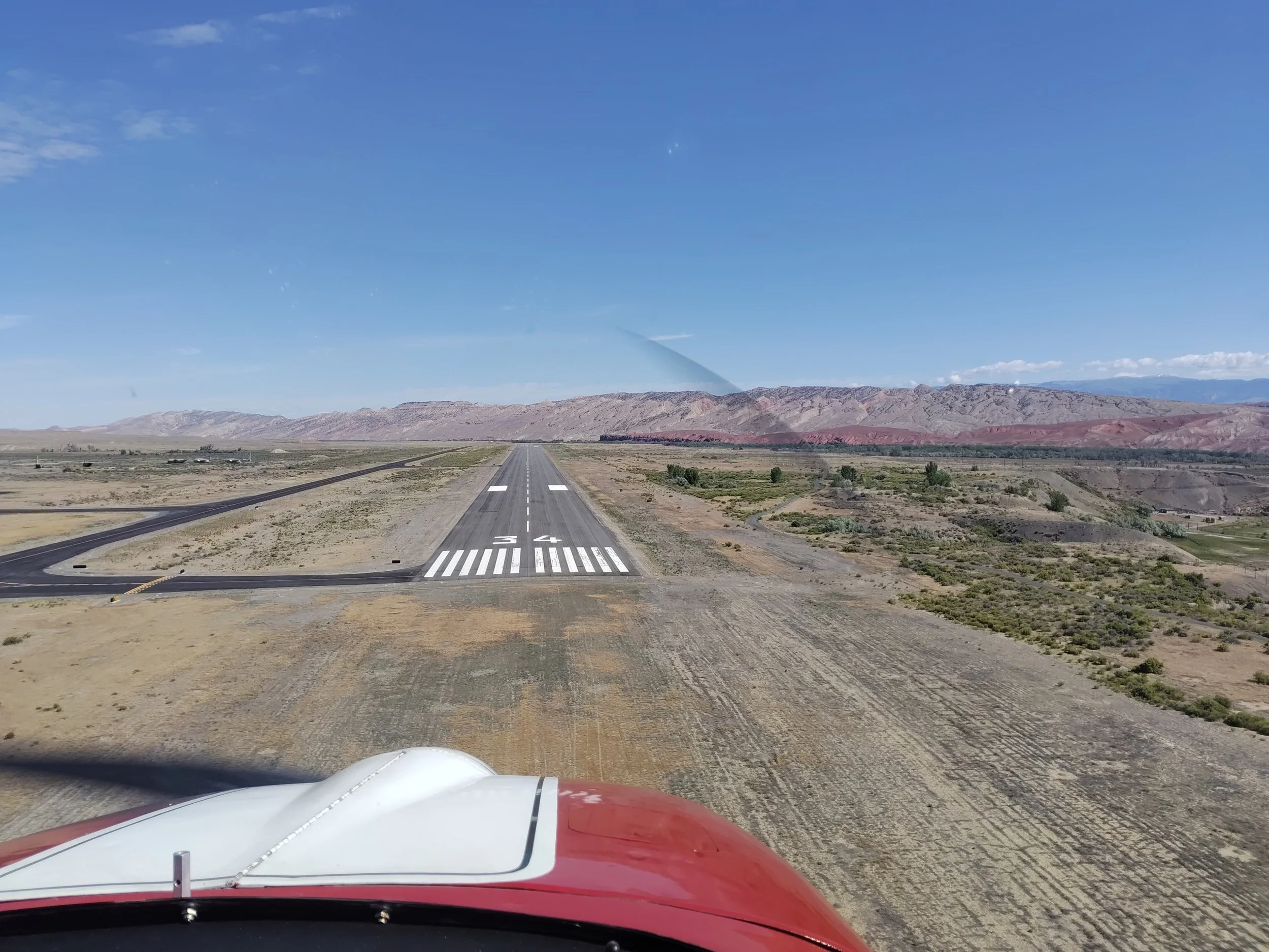 View from inside an airplane cockpit showing a red and white airplane nose on a straight runway in a desert landscape with mountains in the background, under a clear blue sky.