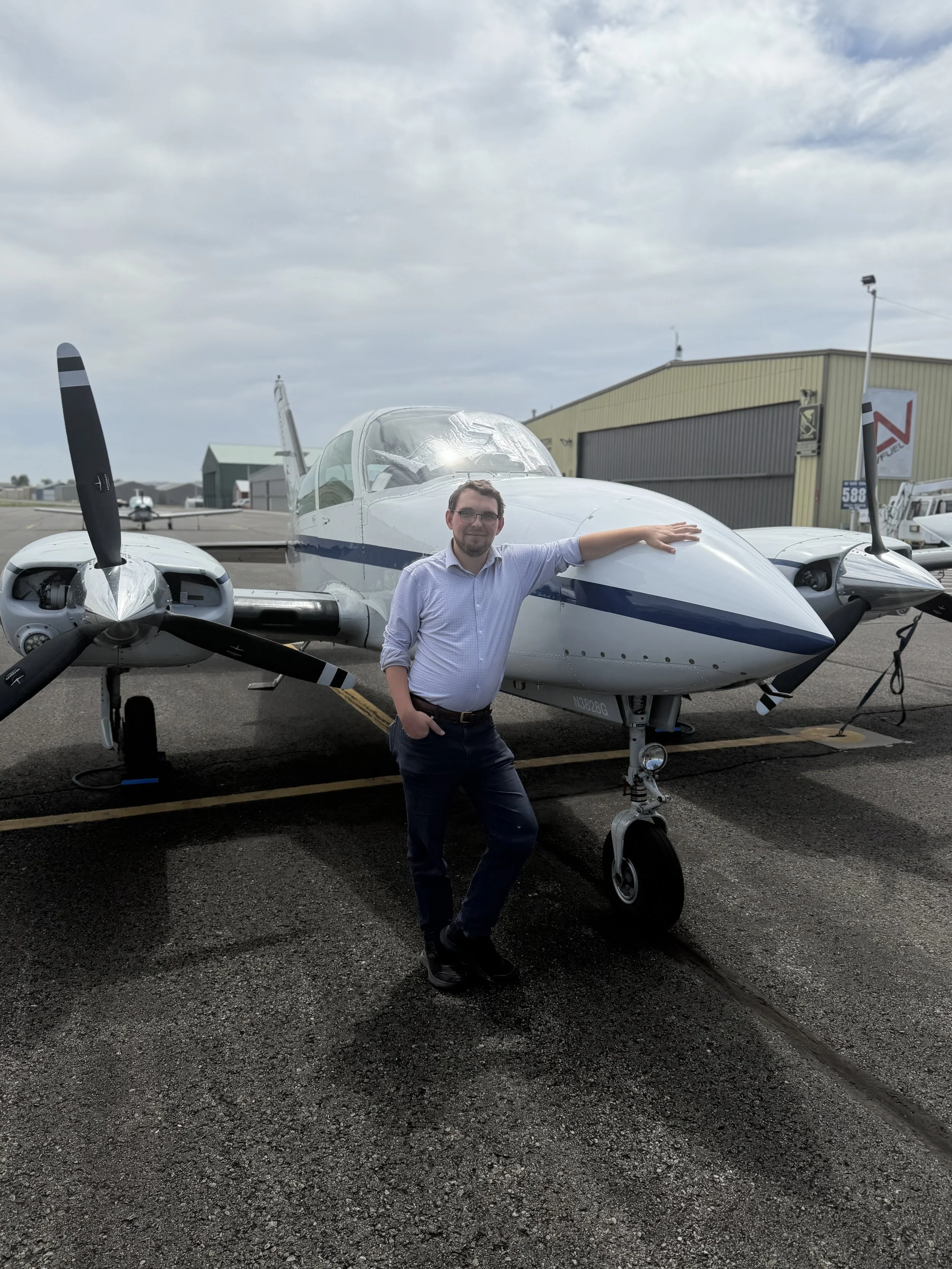 A man standing next to a small aircraft at an airport, smiling, with hangars and other aircraft in the background.