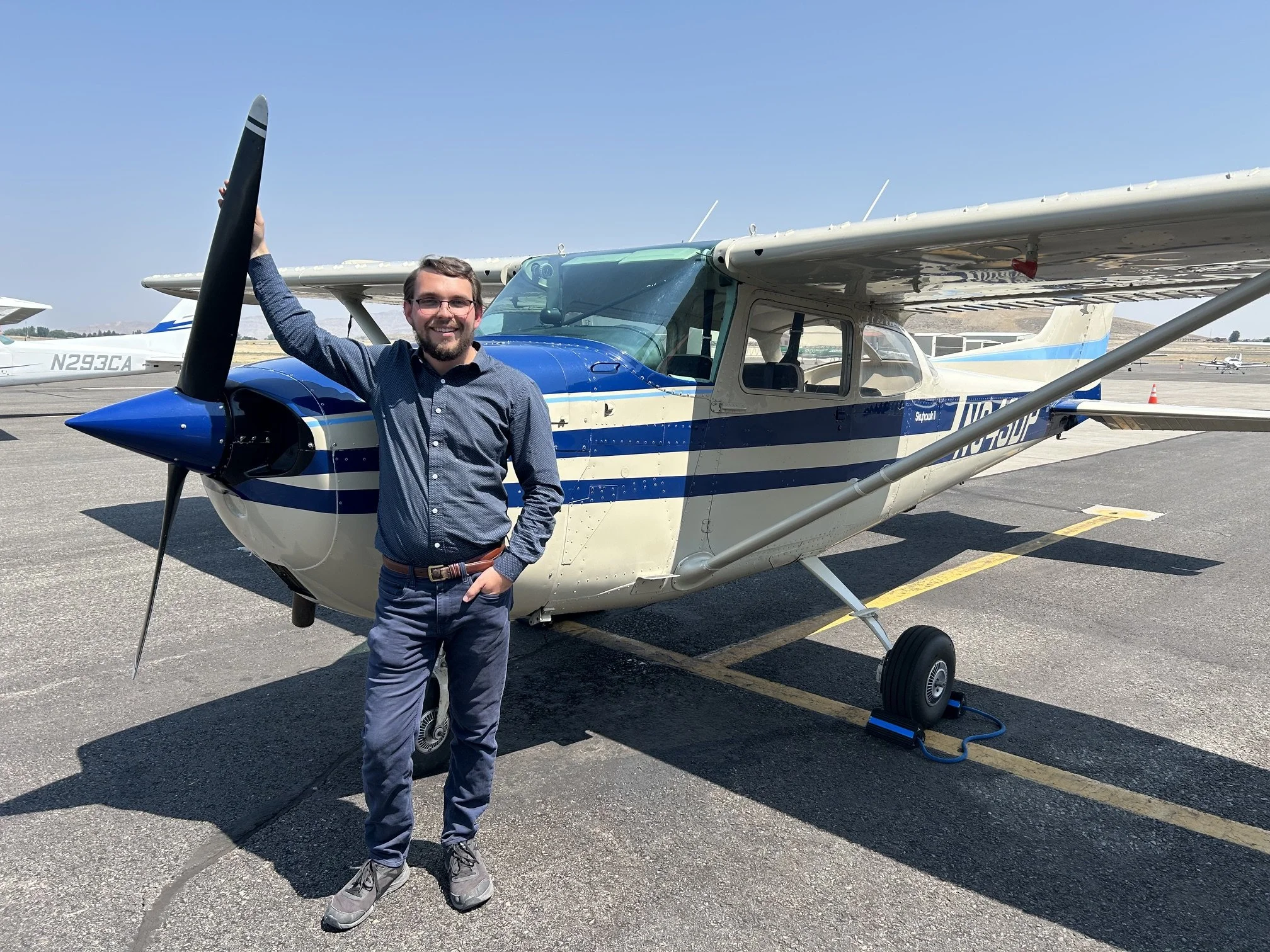 A man standing in front of a small blue and white airplane with a tow cable attached to its nose, at an airfield on a sunny day.