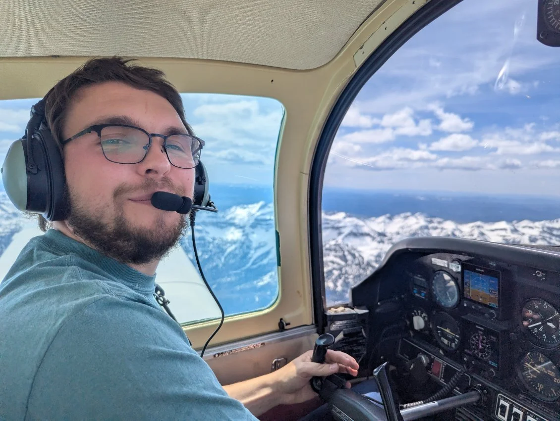 A young man with glasses and a headset smiling while flying an airplane over snow-capped mountains, with a view of the aircraft's cockpit instruments and a blue sky outside the window.