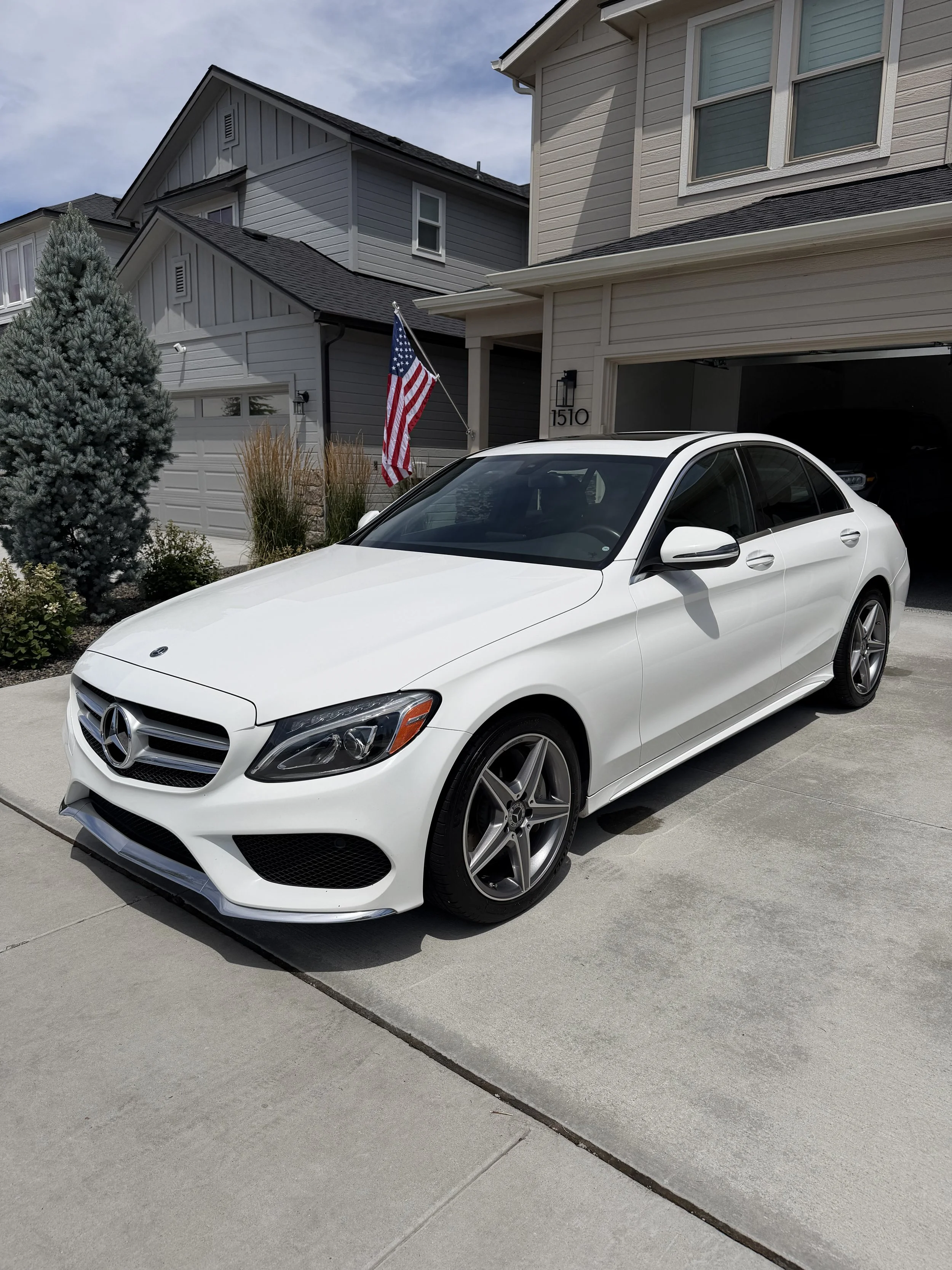 White Mercedes-Benz sedan parked in driveway in front of a house with gray siding, American flag, and garage open.