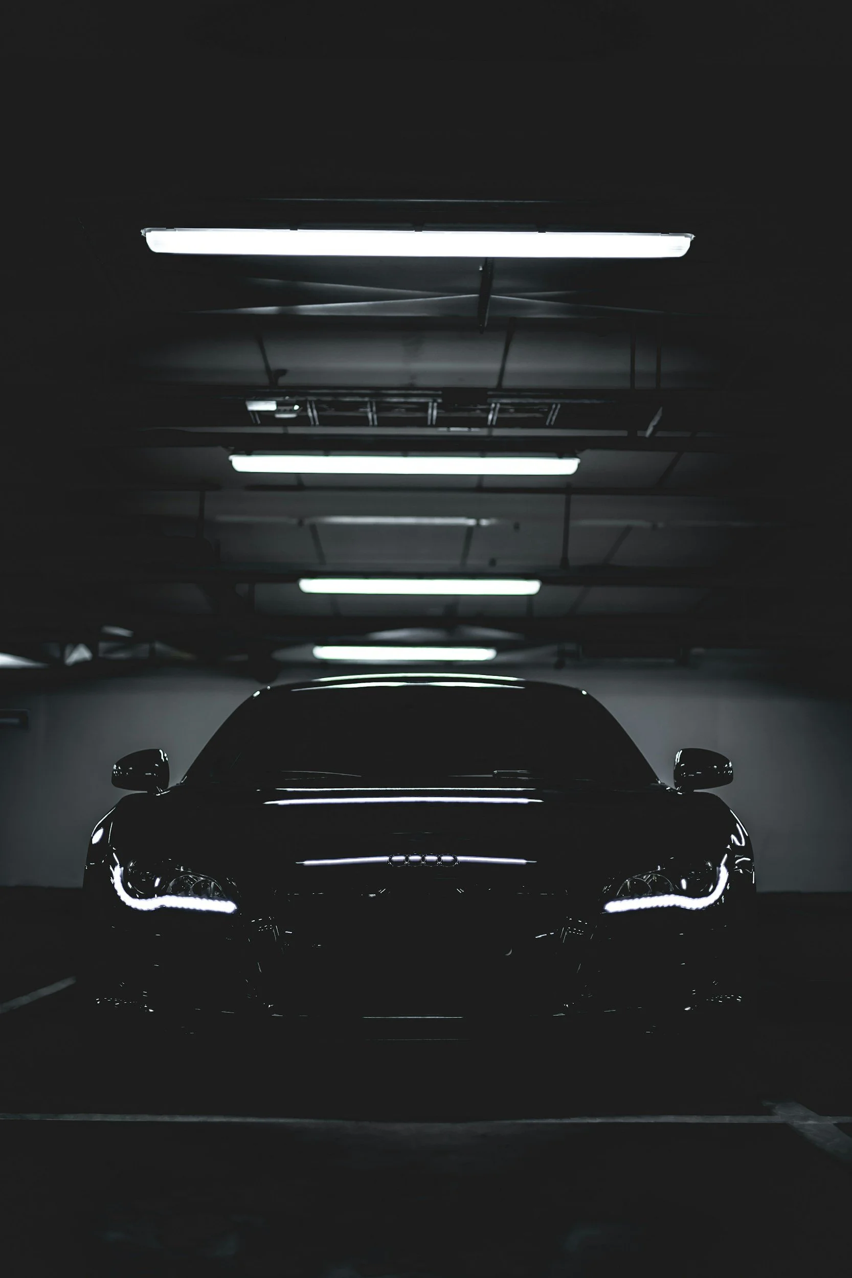 A black luxury car with illuminated headlights parked in a dark underground garage with fluorescent lights on the ceiling.