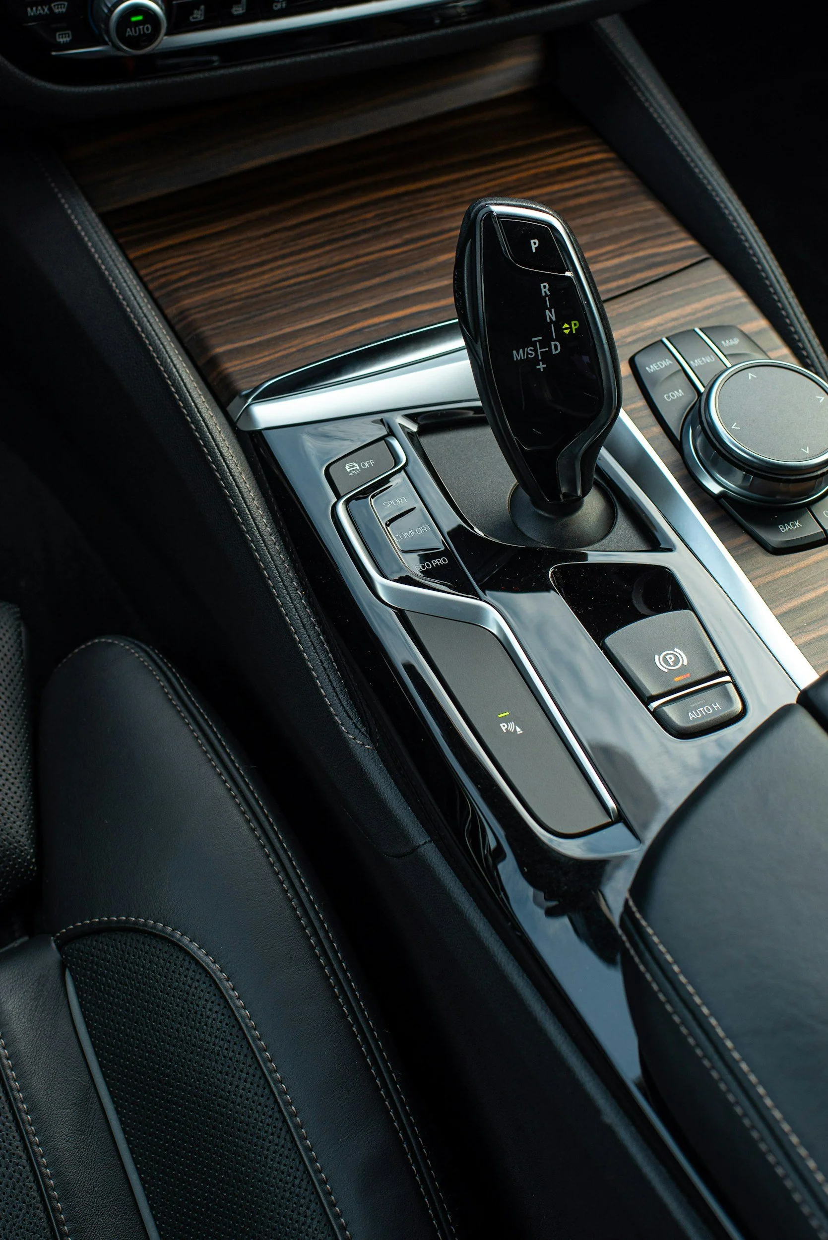 Close-up of a car's gear shift lever situated on a wooden center console with various control buttons and a control wheel nearby.