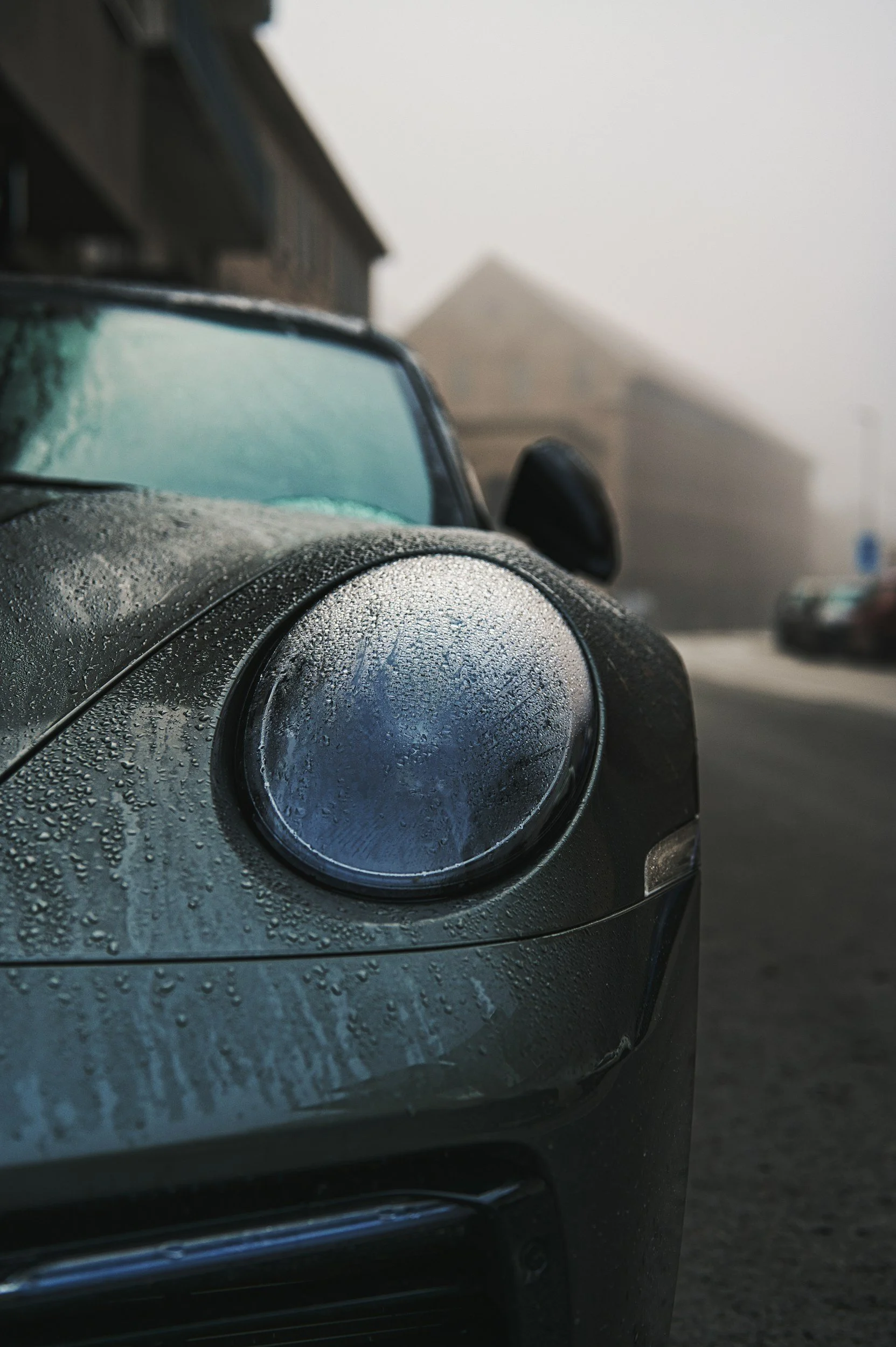Close-up of a black car's front equipped with round headlights, covered in raindrops, with a foggy background containing other vehicles and a building.