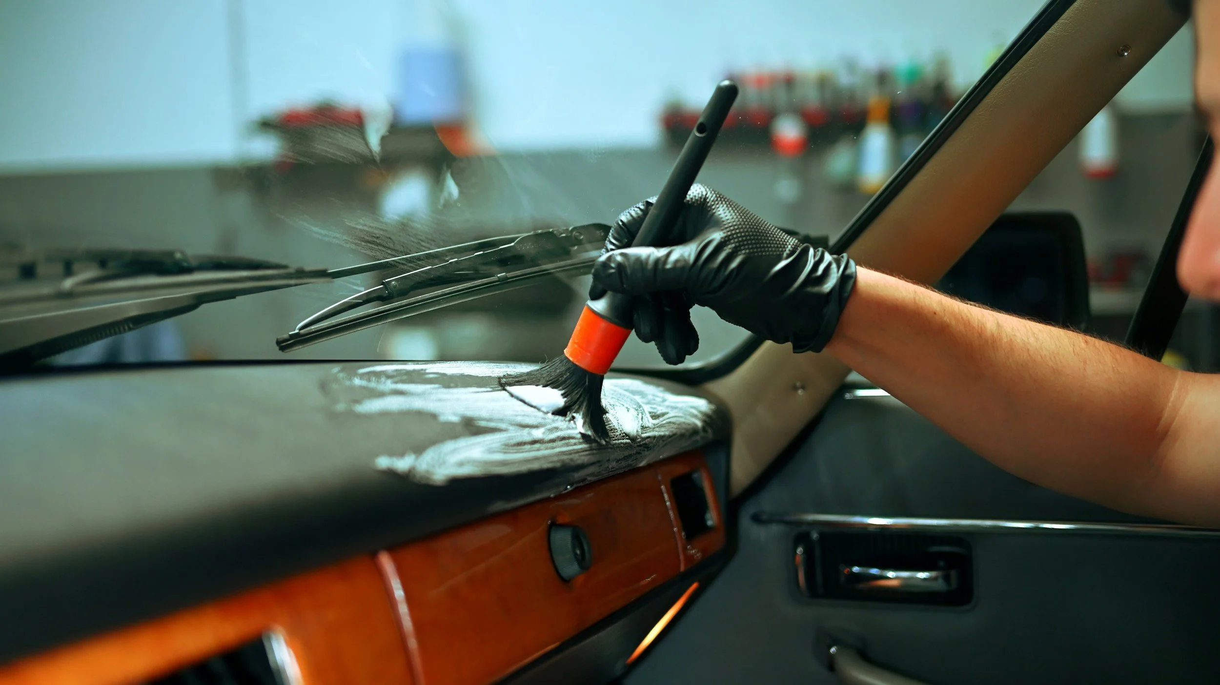 A person wearing a black glove is using a paintbrush to clean or scrub the dashboard of a vehicle, with various bottles and tools blurred in the background.