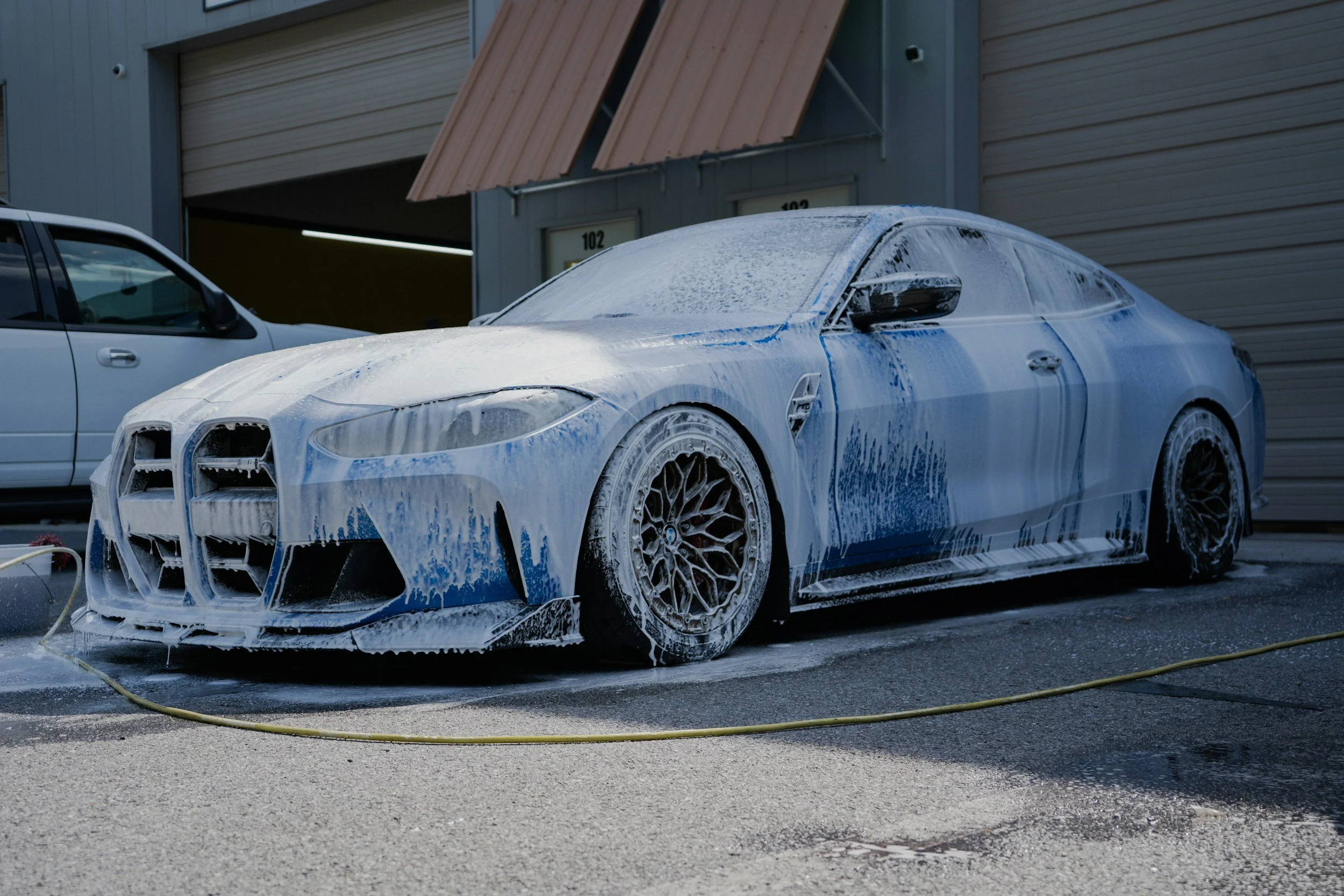 A sports car covered in soap suds during washing, with foam dripping from the hood and wheels, in a parking lot with a building in the background.