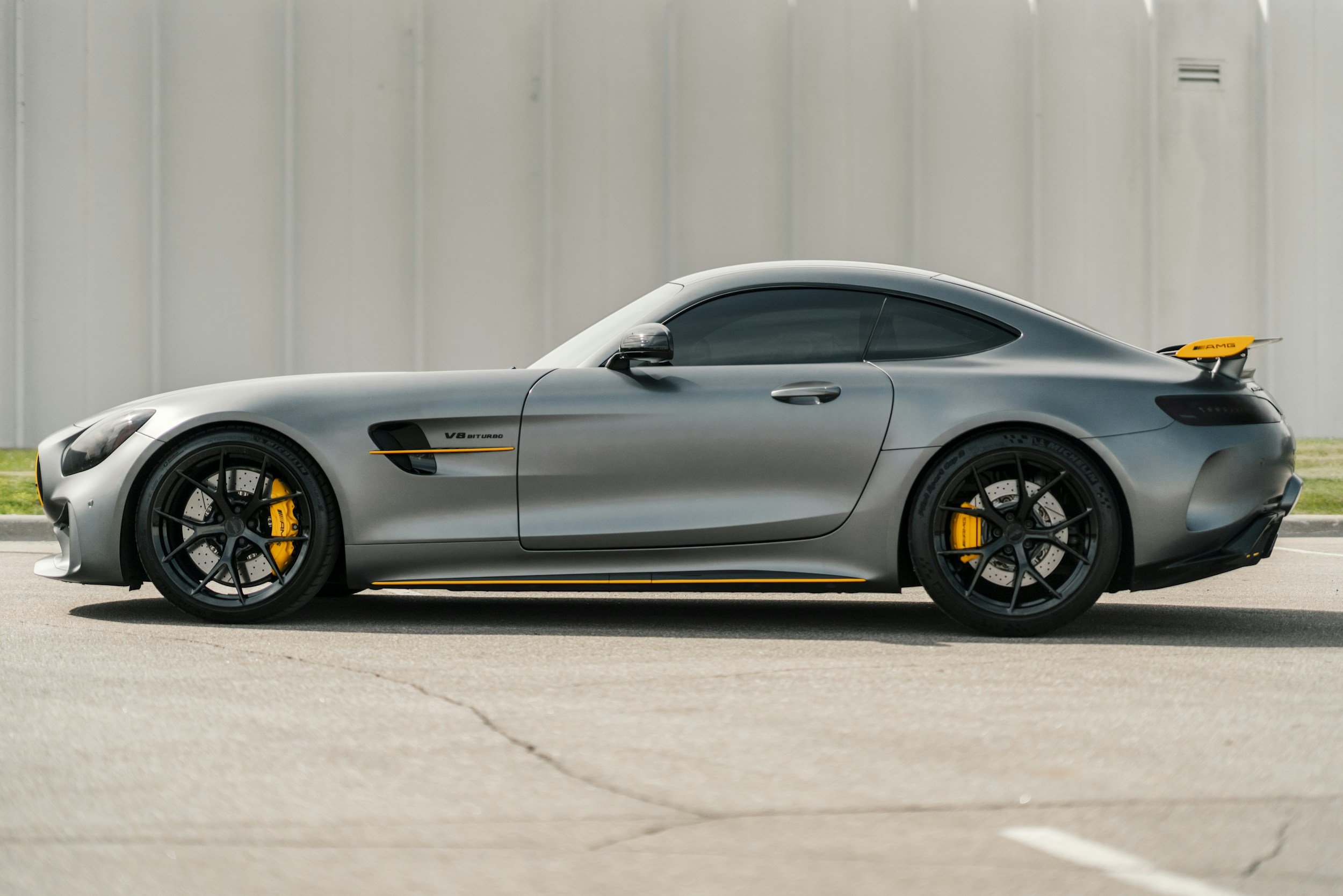 Silver sports car with yellow brake calipers and a yellow rear wing parked on an asphalt surface in front of a gray wall.