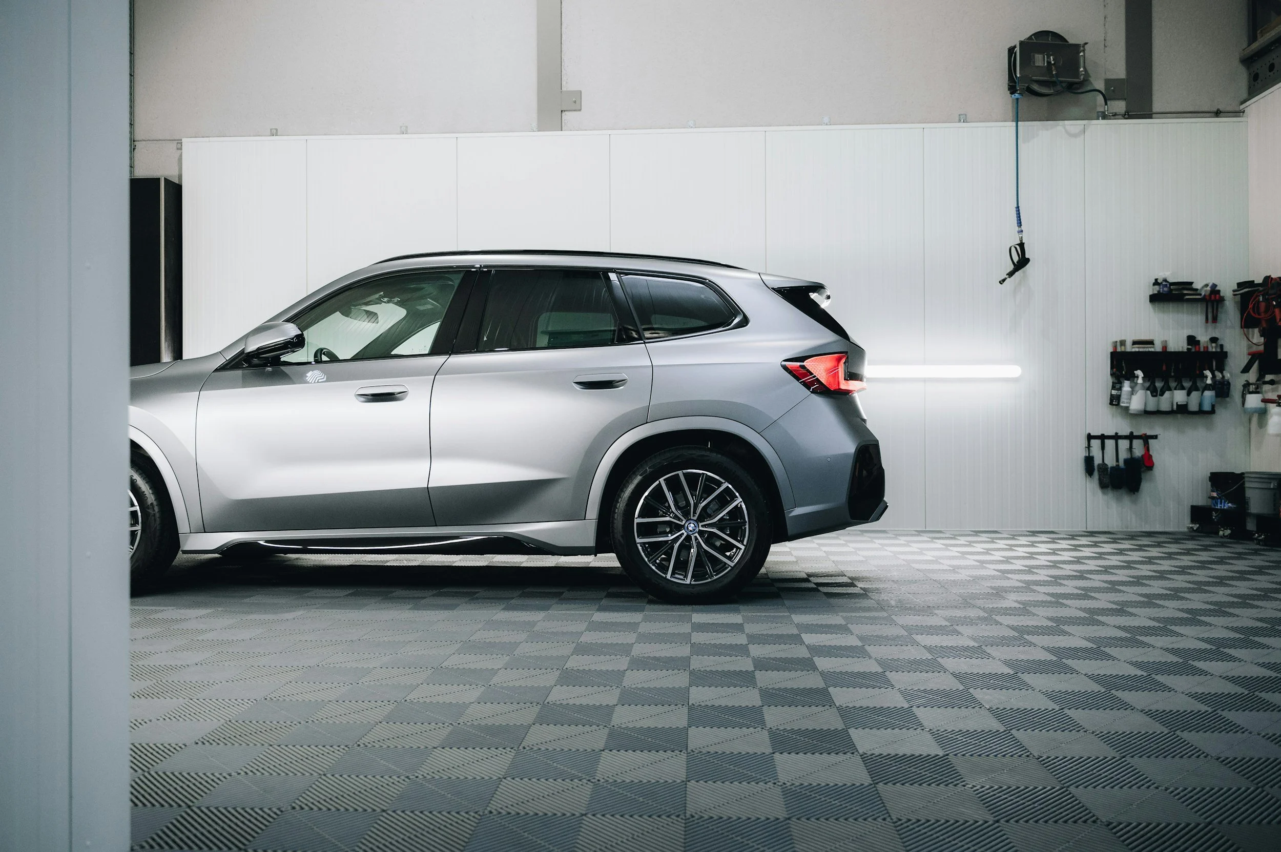 A silver SUV parked in a garage with a gray patterned floor, white wall, and tools organized on shelves.