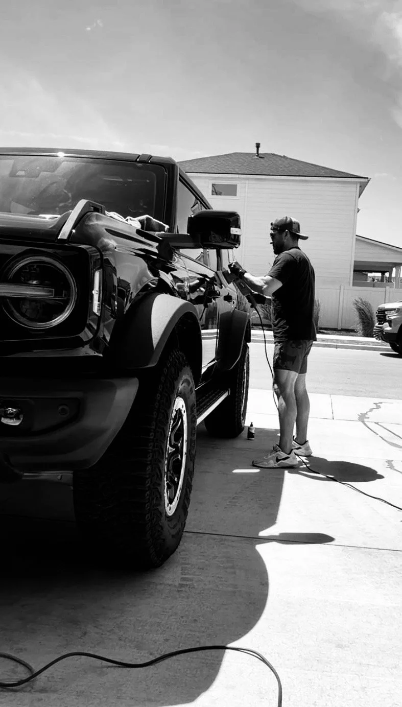 Man polishing a black SUV outside on a driveway in bright sunlight.