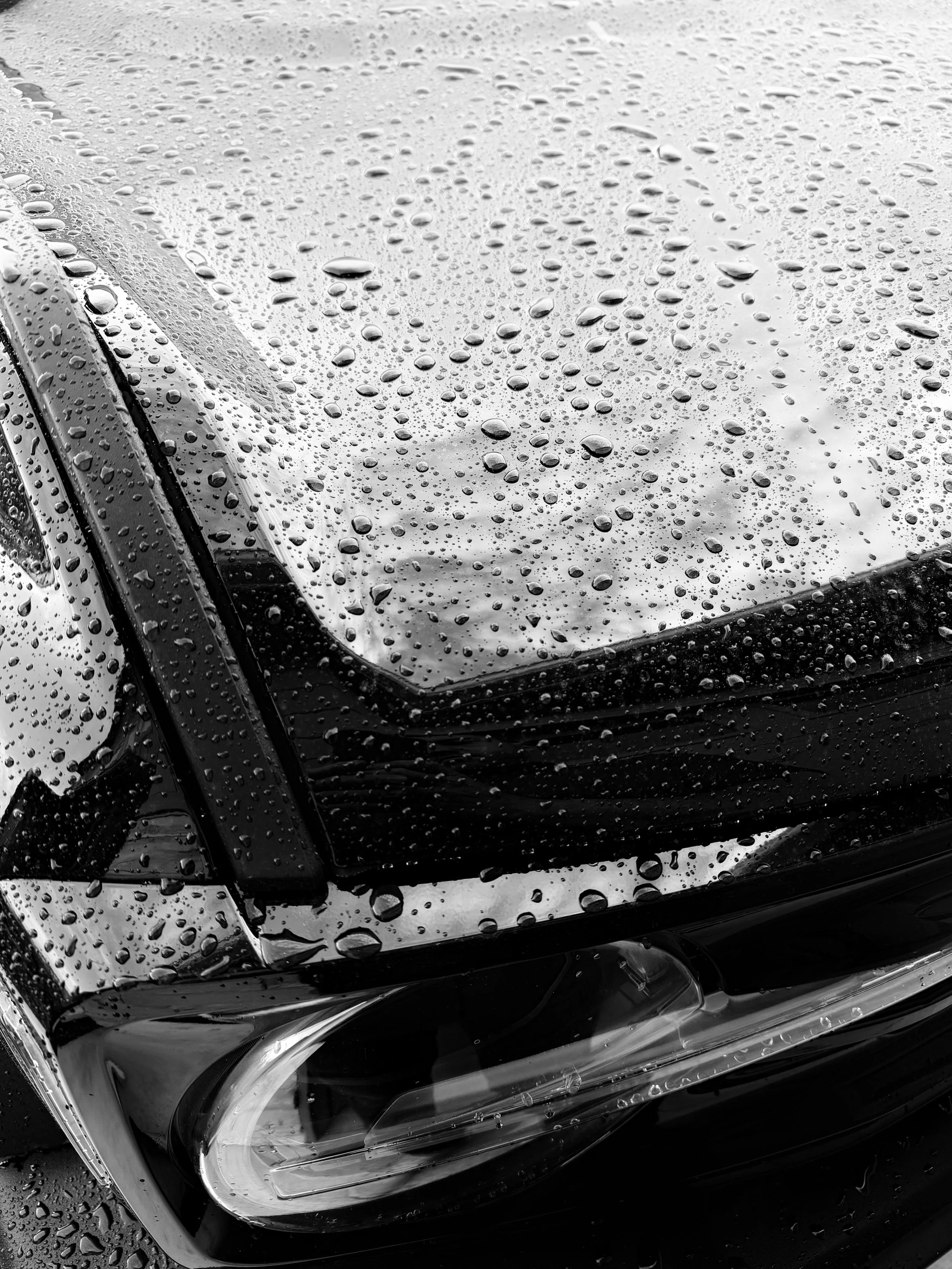 Close-up of a black car's front corner and headlight with water droplets on the surface, rain-soaked scene.